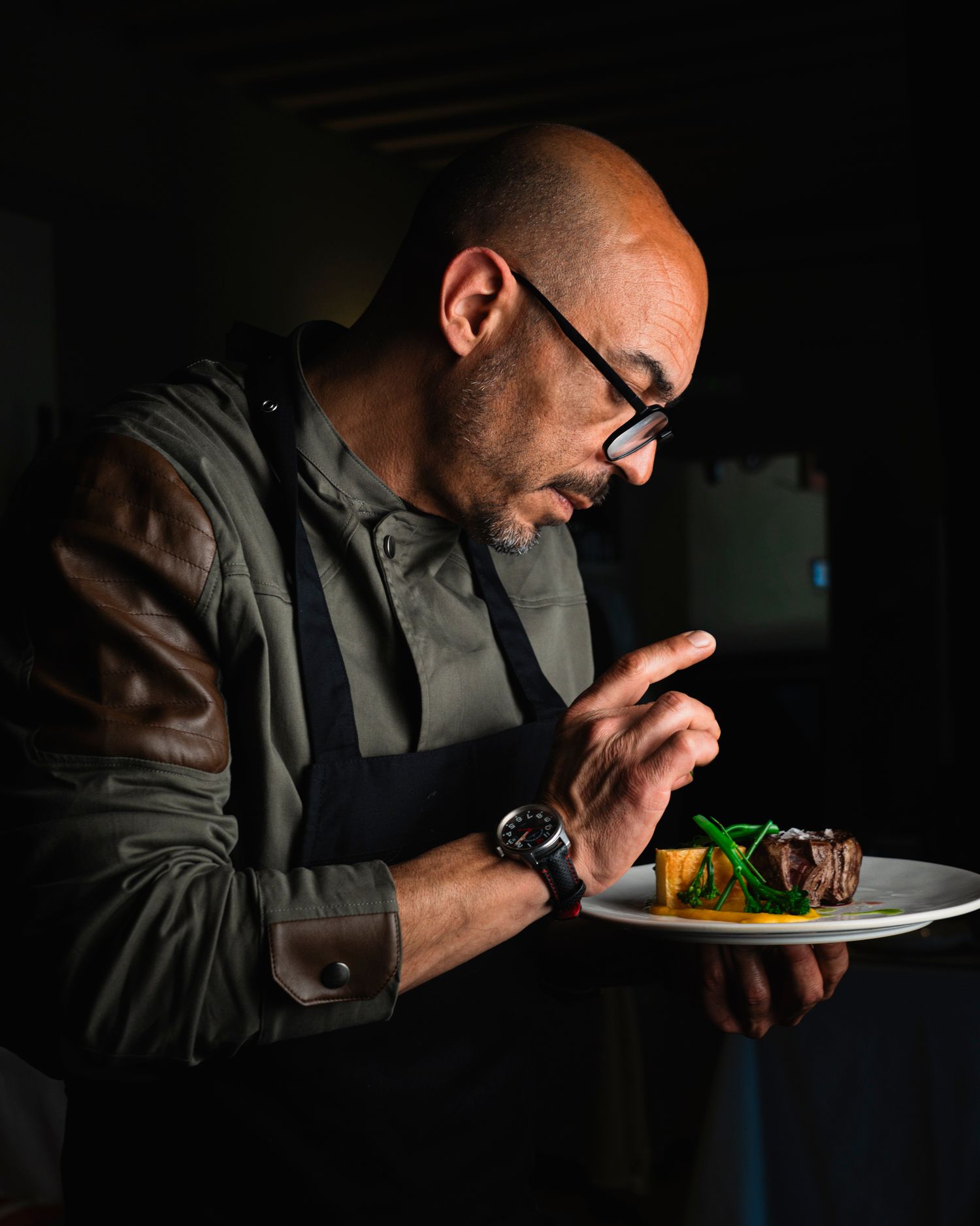 Bald chef with glasses carefully plating a gourmet dish in a dark professional kitchen.