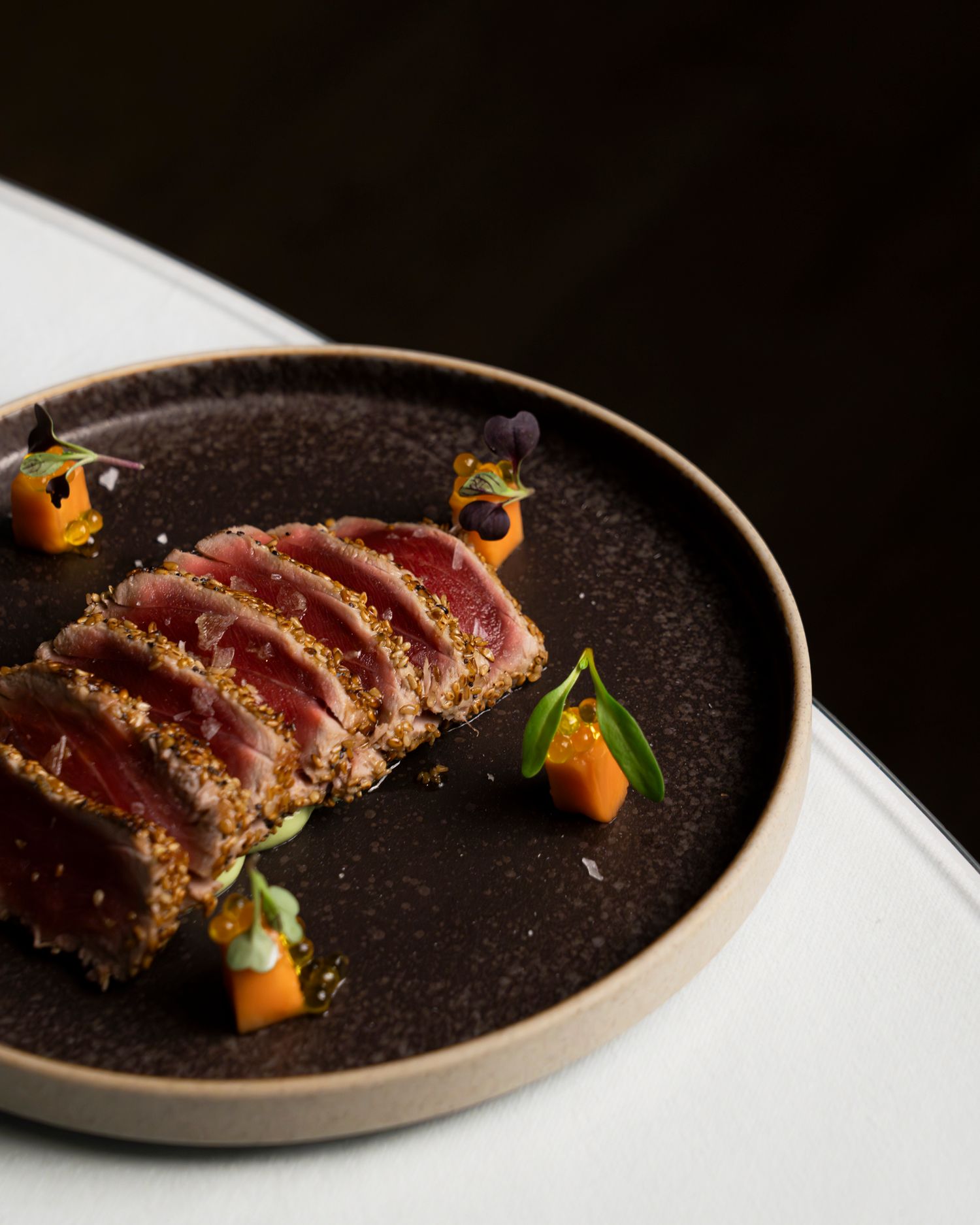 Seared sesame-crusted tuna slices on a dark ceramic plate, garnished with mango cubes and edible flowers.