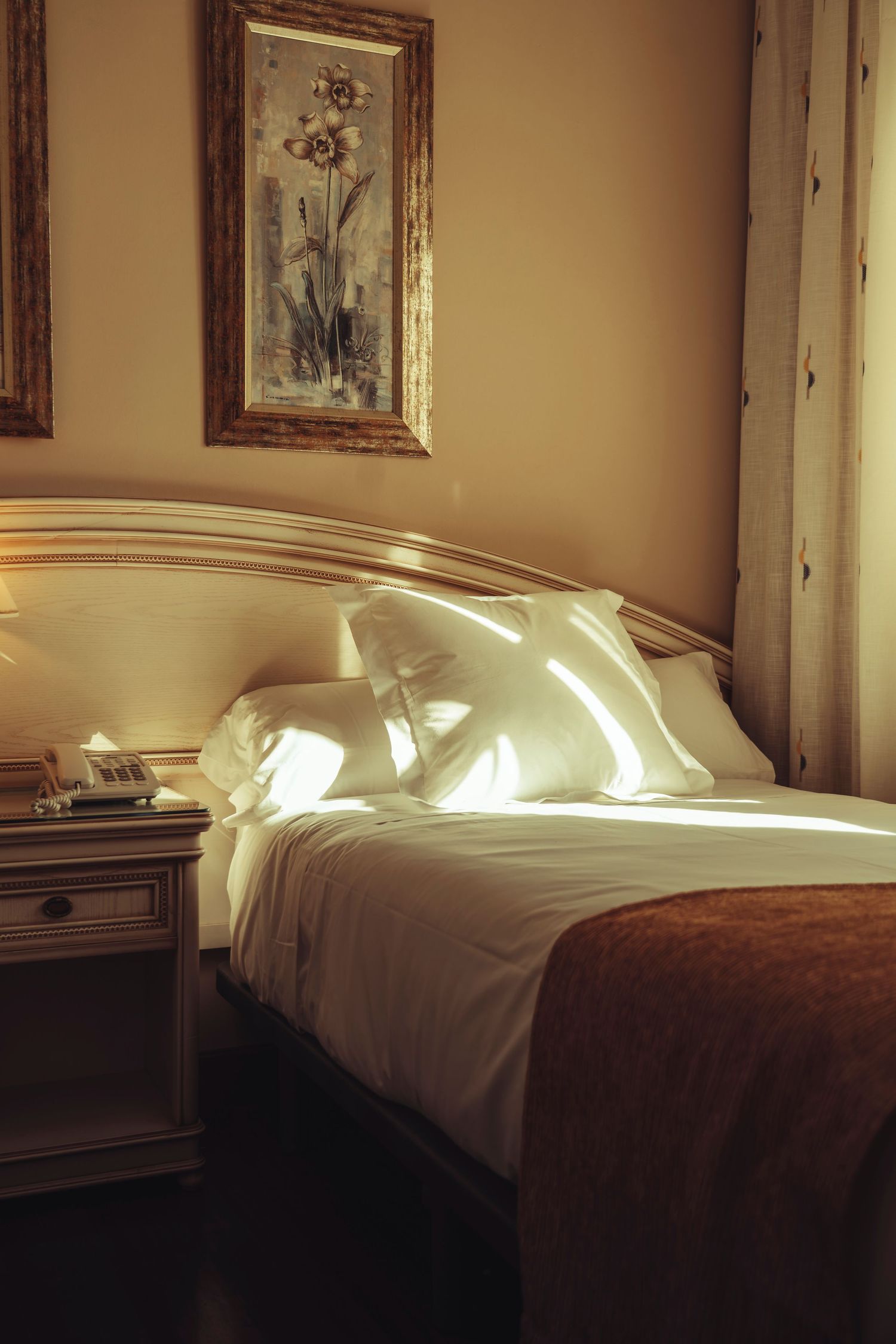 Elegant hotel room with white upholstered headboard, crisp white pillows, brown throw, and framed floral artwork above bed.