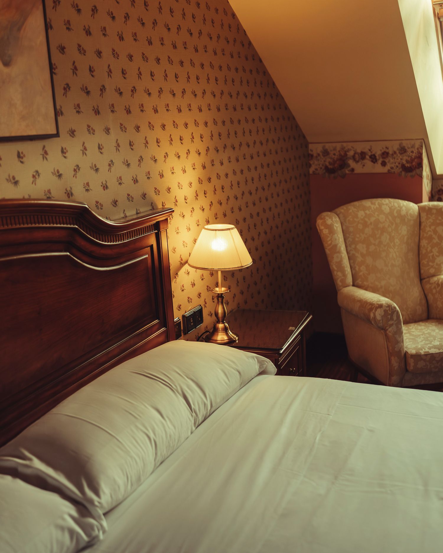 Cozy hotel bedroom with floral wallpaper, wooden headboard, bedside lamp, and armchair under sloped ceiling.