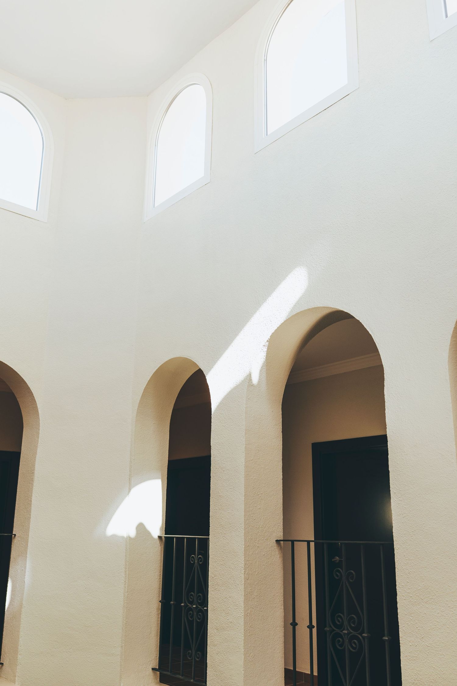 White stucco building interior with arched doorways, wrought iron railings, and tall arched windows casting natural light on walls.