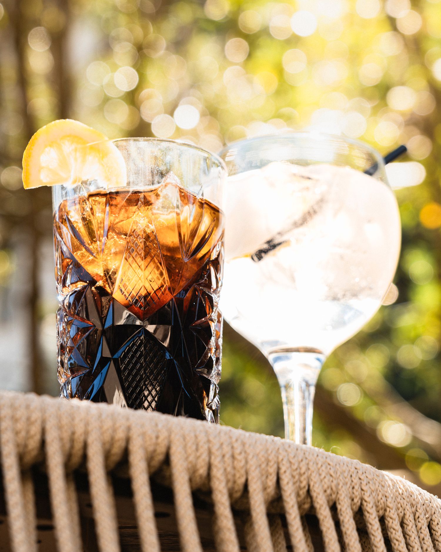 Two cocktails in crystal glasses with lemon garnish on a wicker surface with bokeh background.