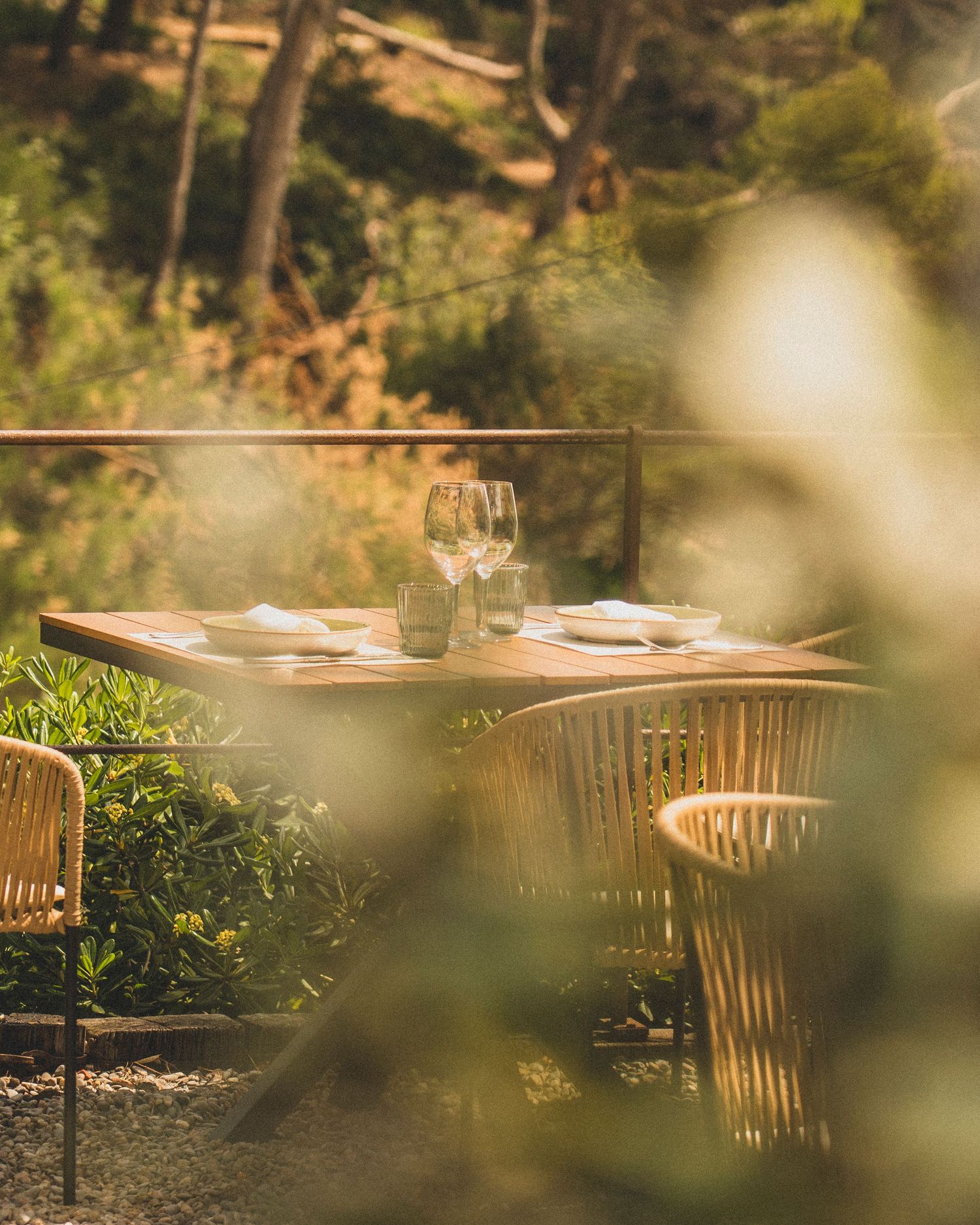 Sunlit outdoor restaurant table set with wine glasses and plates surrounded by lush Mediterranean garden greenery.