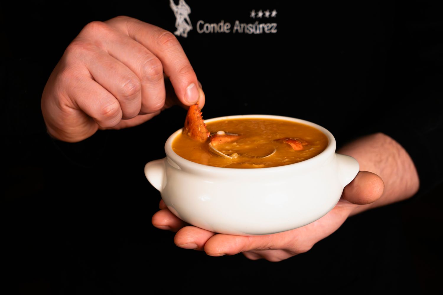 Chef holding a white bowl of rich seafood soup with a shrimp garnish at Conde Ansarez.