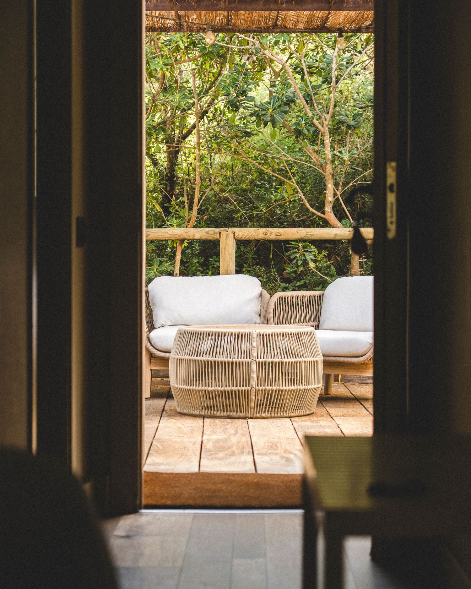 View through open door to a serene outdoor patio with rattan furniture and lush greenery.