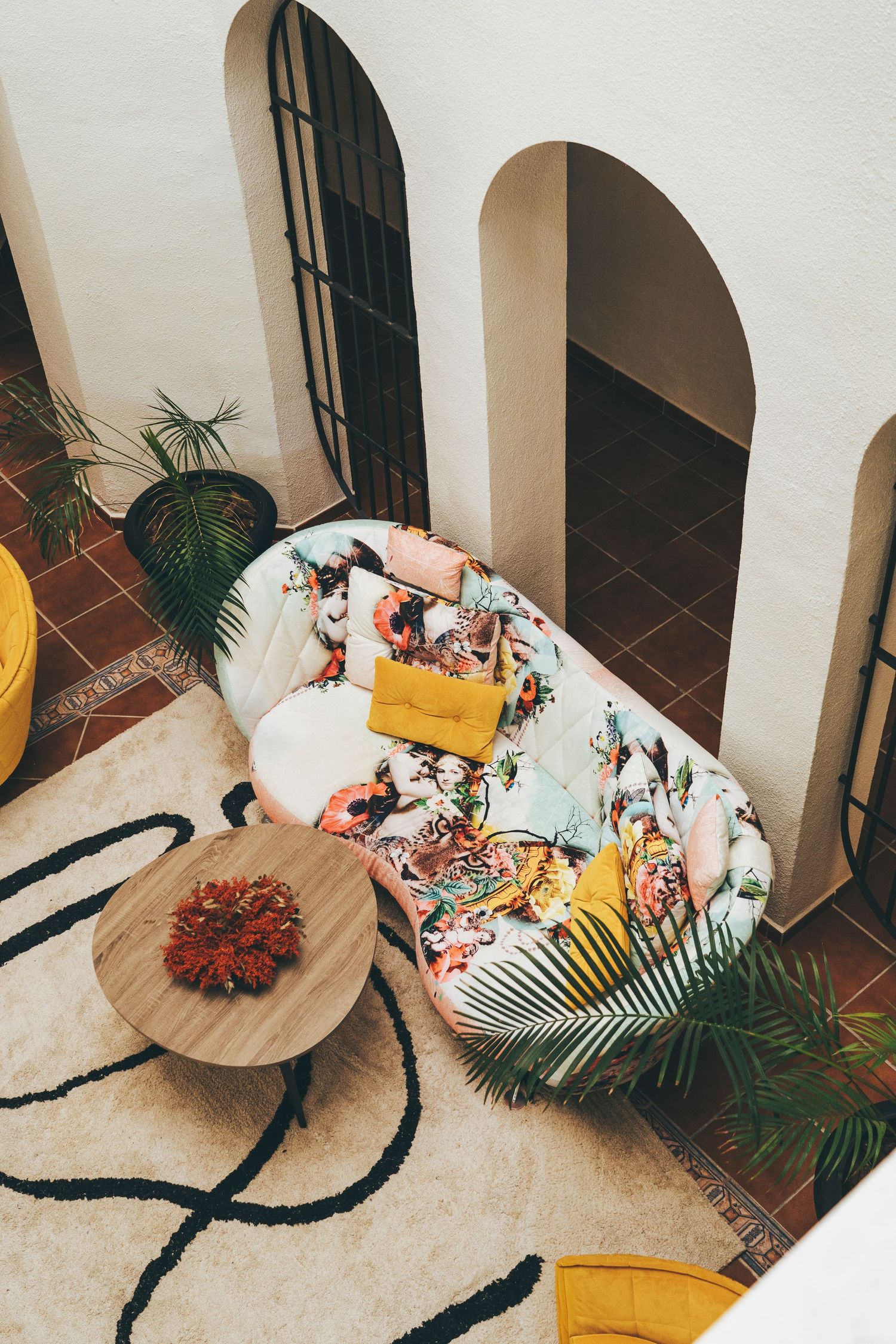 Aerial view of a colorful floral sofa with yellow cushions on a patterned rug beside a round wooden coffee table.
