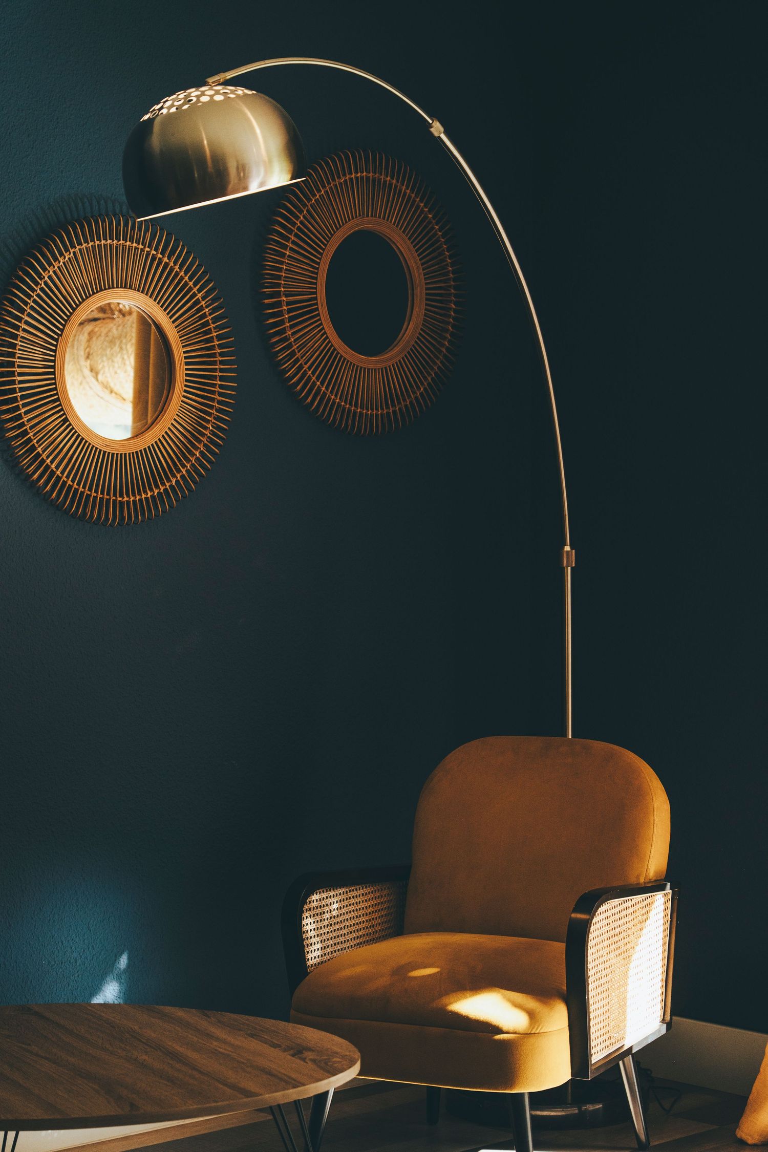 Mid-century modern living room with gold arc floor lamp, velvet mustard armchair, and sunburst mirrors on dark teal wall.