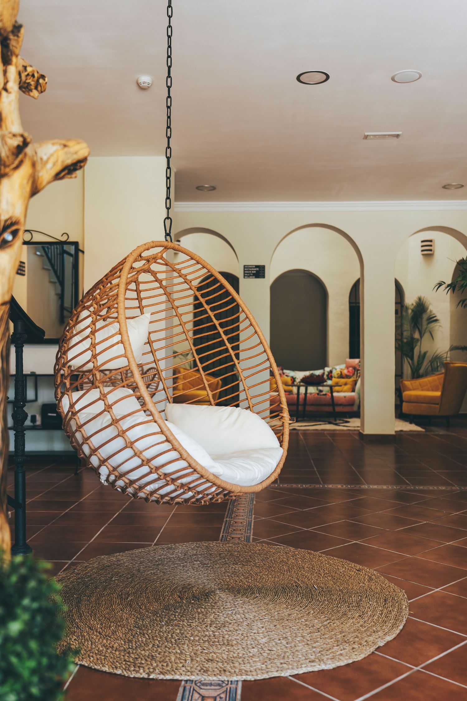Bohemian rattan hanging egg chair with white cushion suspended from ceiling over round woven rug in elegant hotel lobby.