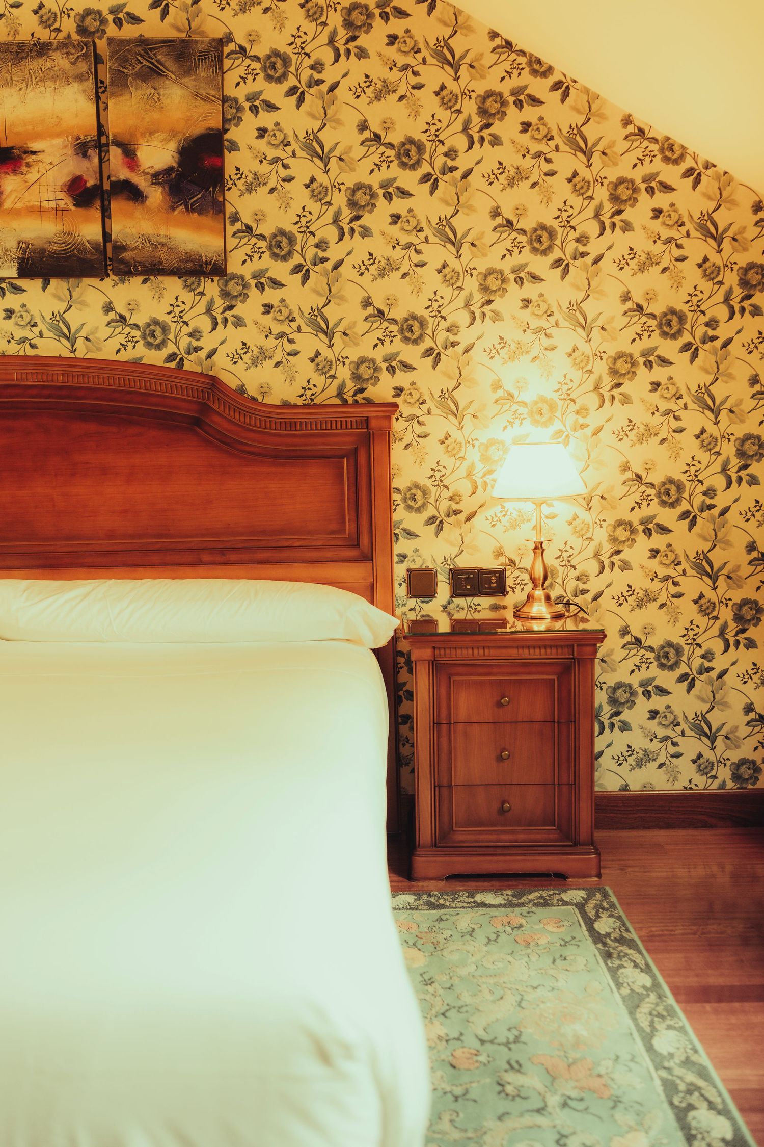 Classic hotel bedroom with ornate floral wallpaper, wooden headboard, white bedding, and vintage bedside lamp on nightstand.