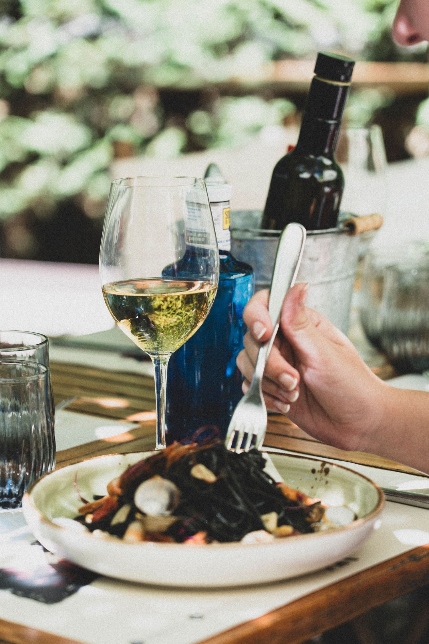 Hand holding a fork over black squid ink pasta beside a glass of white wine outdoors.