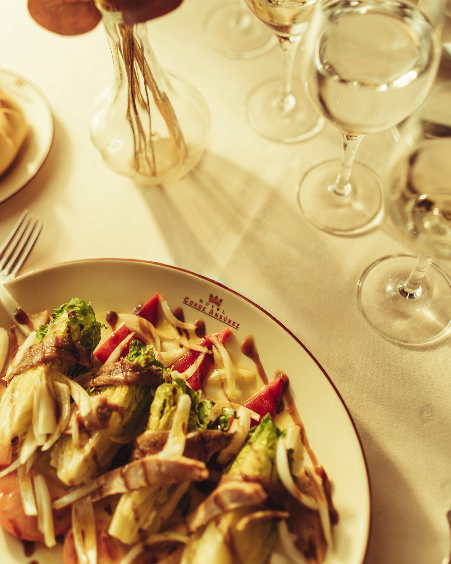Fresh vegetable salad on a branded restaurant plate beside elegant wine glasses on white linen.