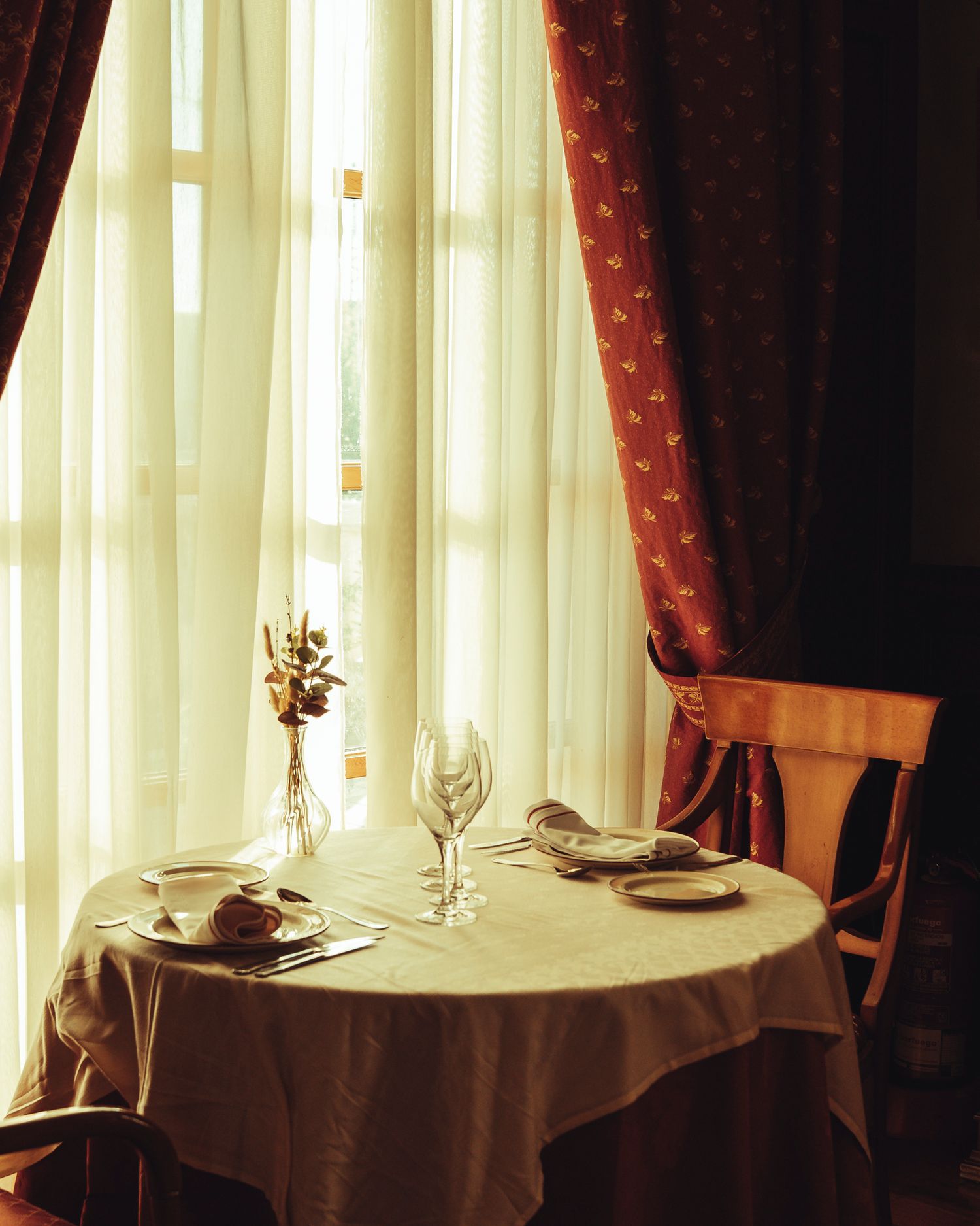 Elegant restaurant table for two set with wine glasses near sunlit sheer curtains and red drapes.
