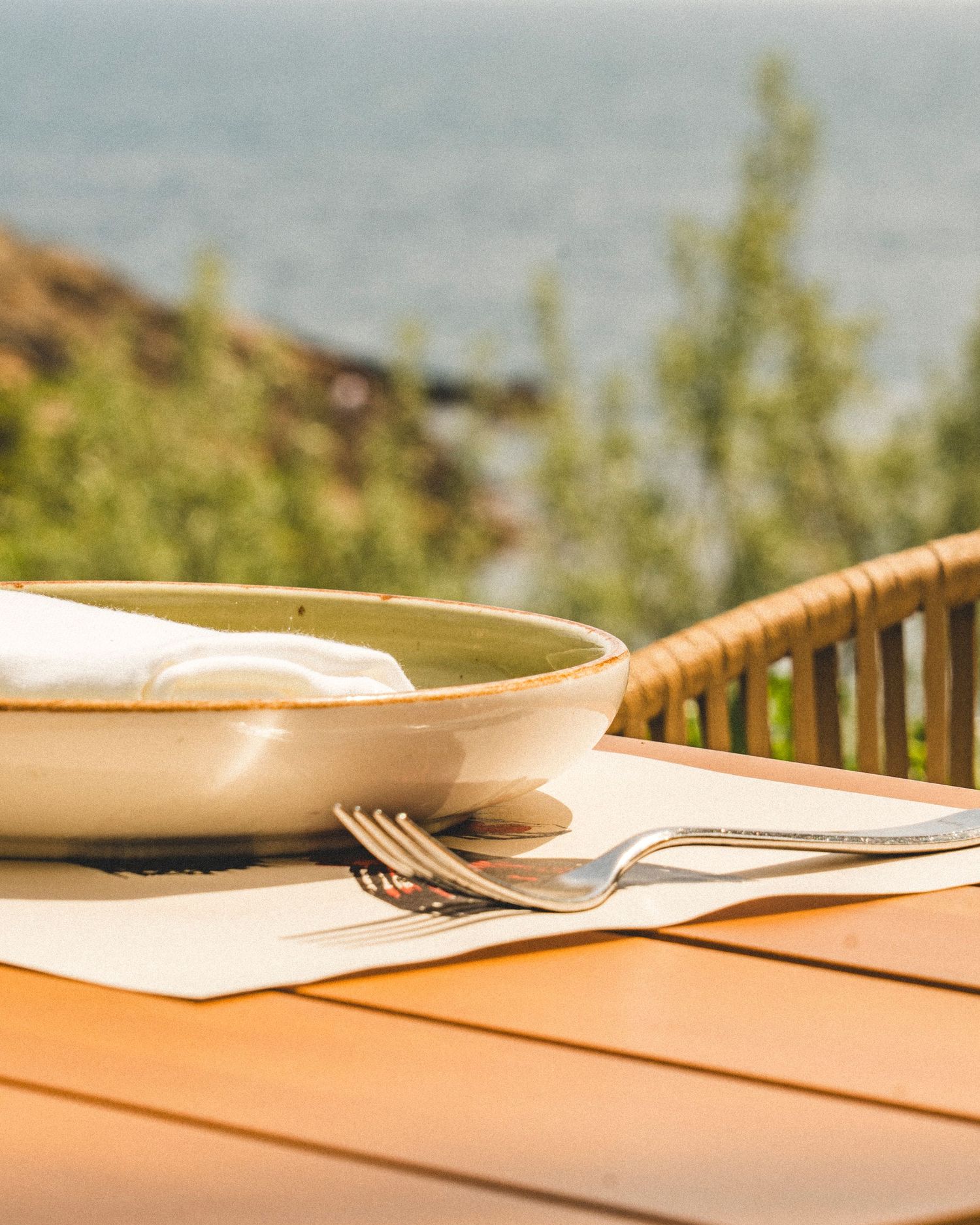 Rustic ceramic bowl with silverware on a white placemat at an outdoor seaside restaurant table.