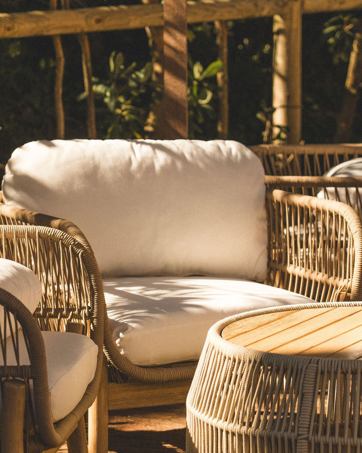 Outdoor patio with natural rattan lounge chairs, white cushions, and wooden pergola surrounded by lush greenery.