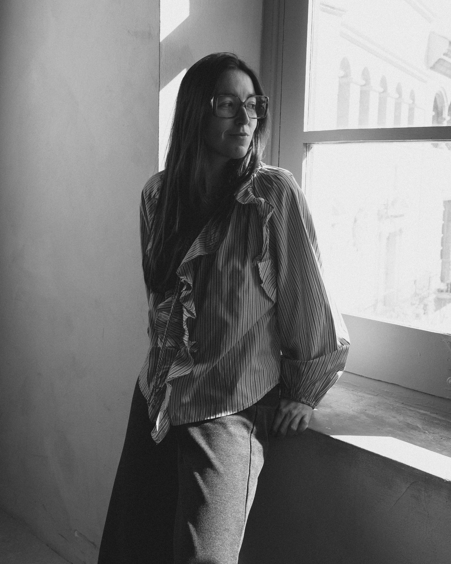 Young woman with long hair and glasses posing by a window in a striped blouse, black and white photo.