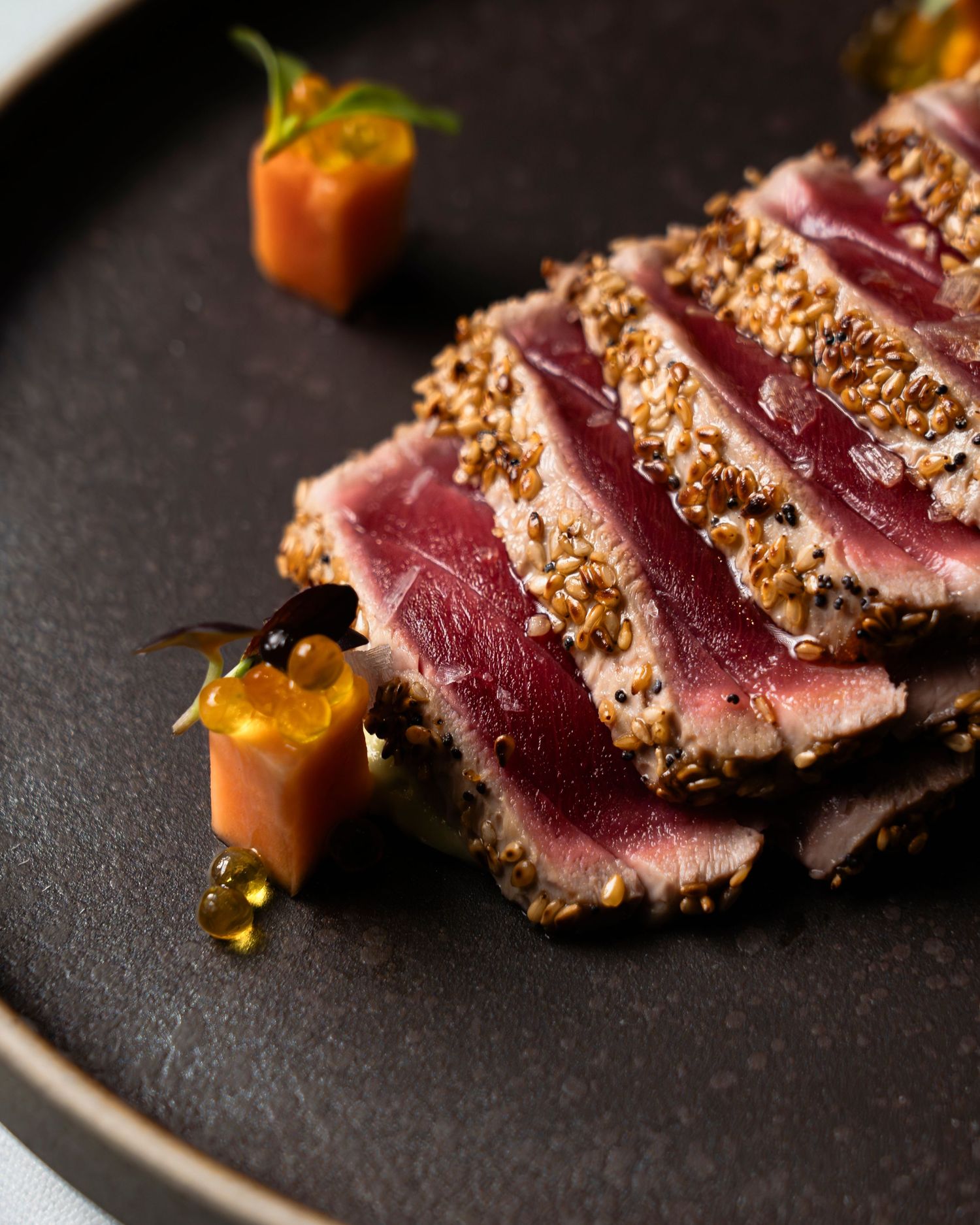 Close-up of sesame-crusted seared tuna slices with vibrant orange garnish on a dark plate.