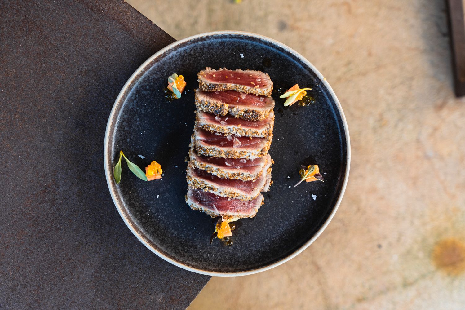 Seared tuna steak sliced and plated on a dark ceramic dish with edible flowers and sesame crust.