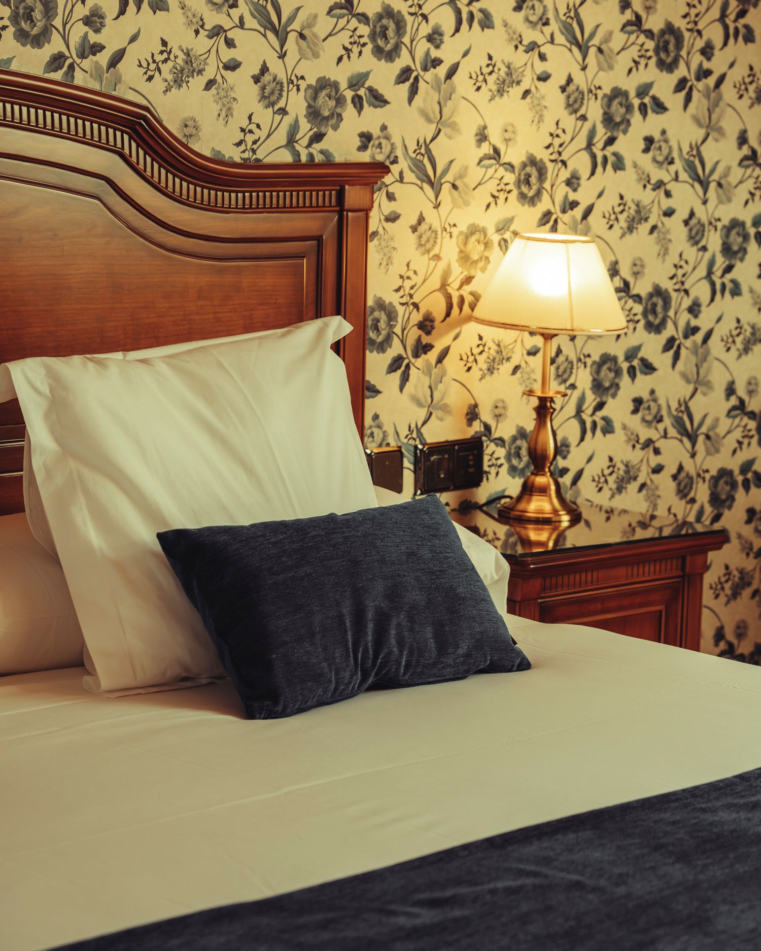 Classic hotel bed with wooden headboard, white pillows, navy cushion, and glowing lamp against floral wallpaper.