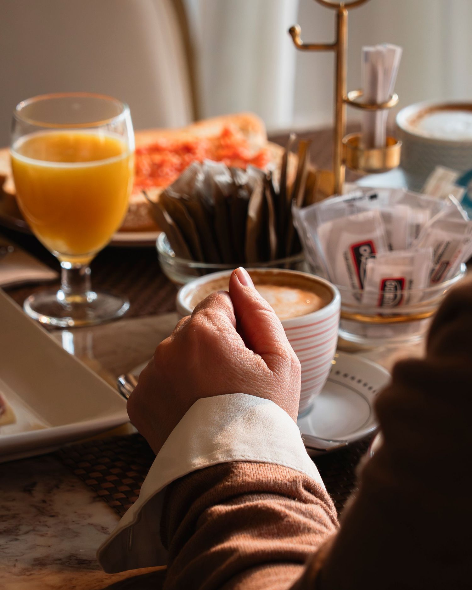 Person holding a latte cup at a breakfast table with orange juice, toast, and sugar packets.