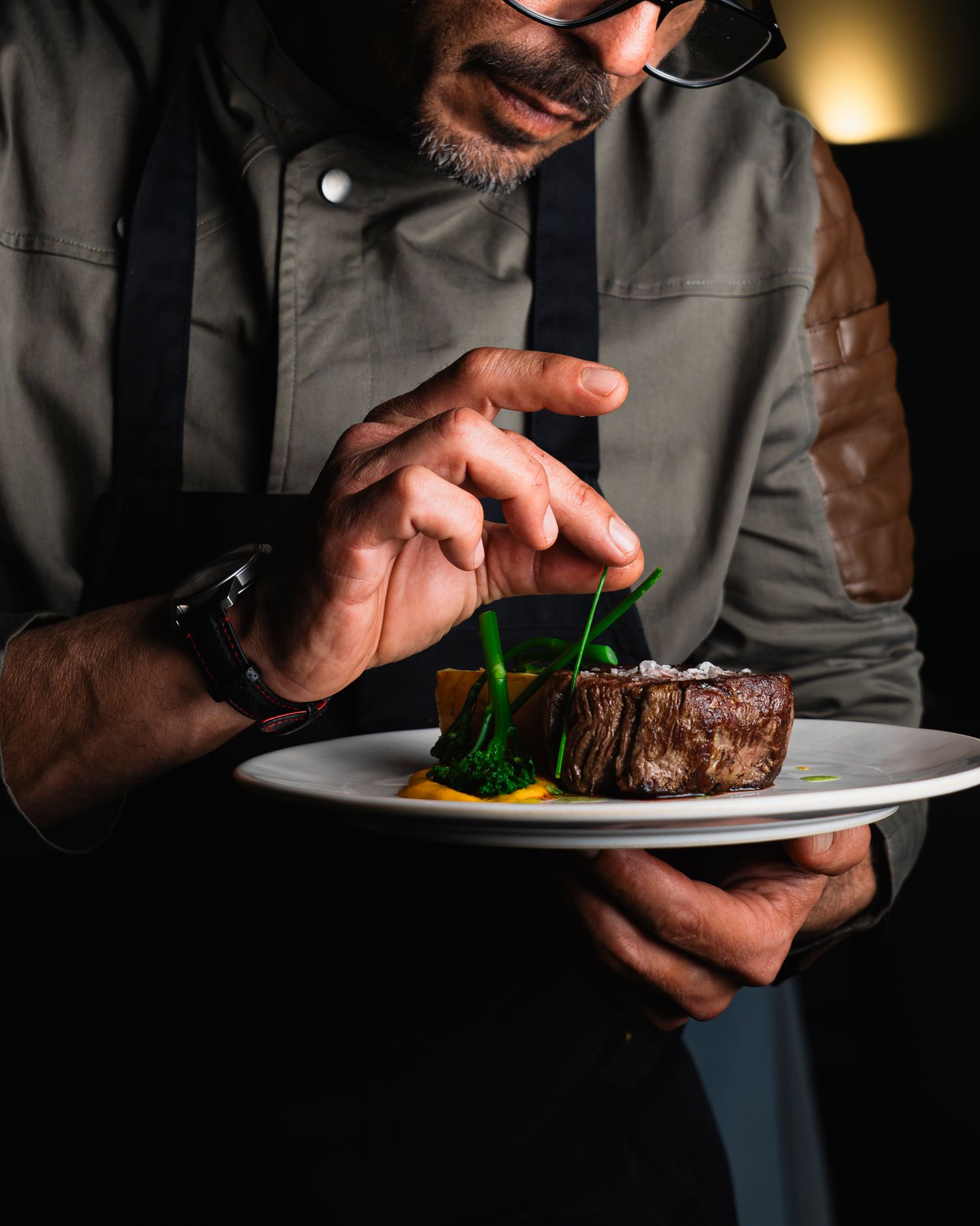 Chef carefully garnishes a seared steak on a white plate, wearing a gray jacket and watch in a dark restaurant setting.