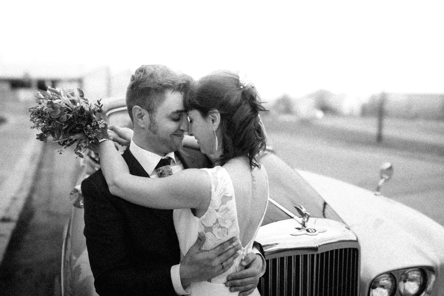 Black and white photo of a bride and groom sharing a romantic moment beside a classic white car on their wedding day.