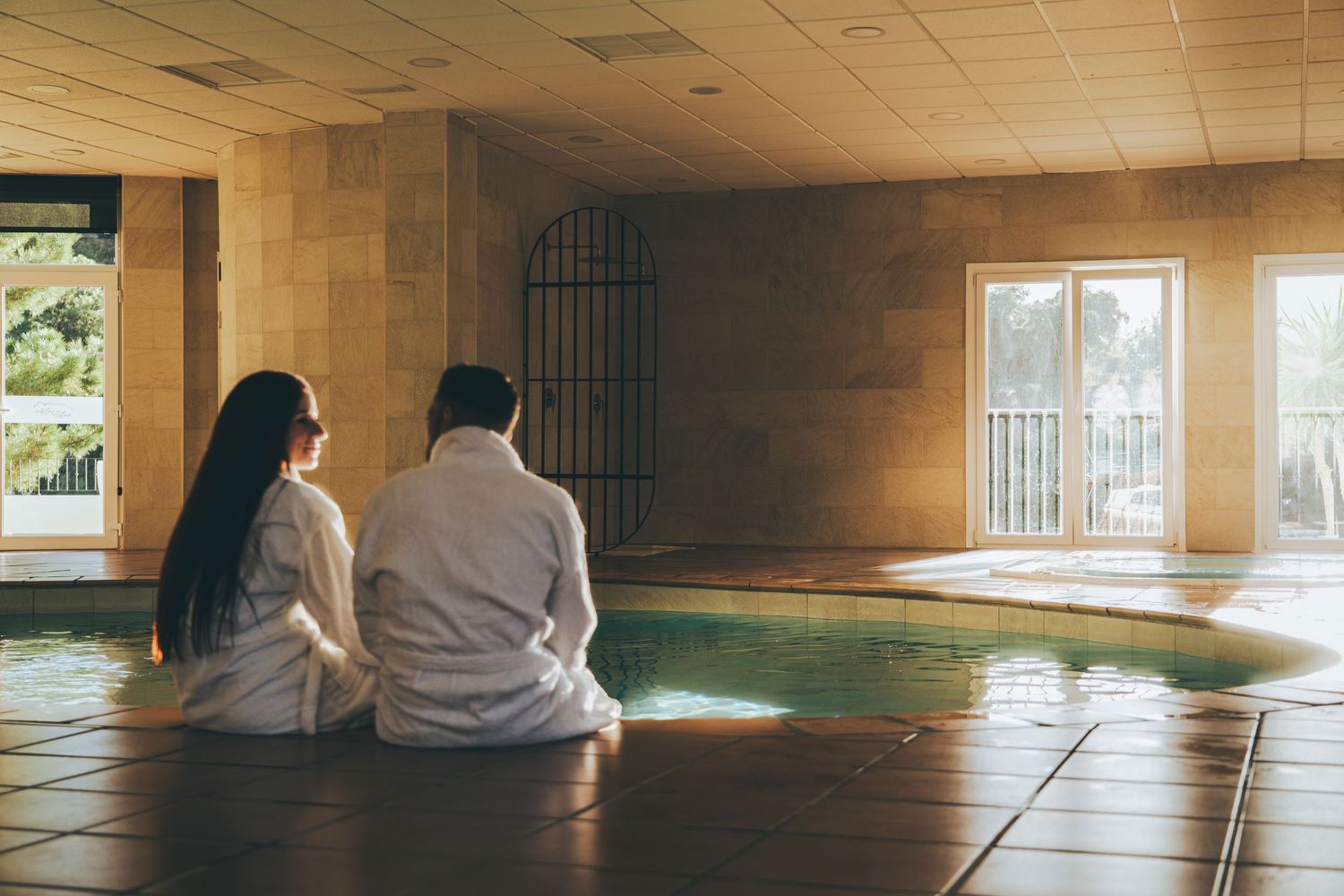 Couple in white robes sitting poolside in an elegant indoor spa with warm stone walls and natural light.