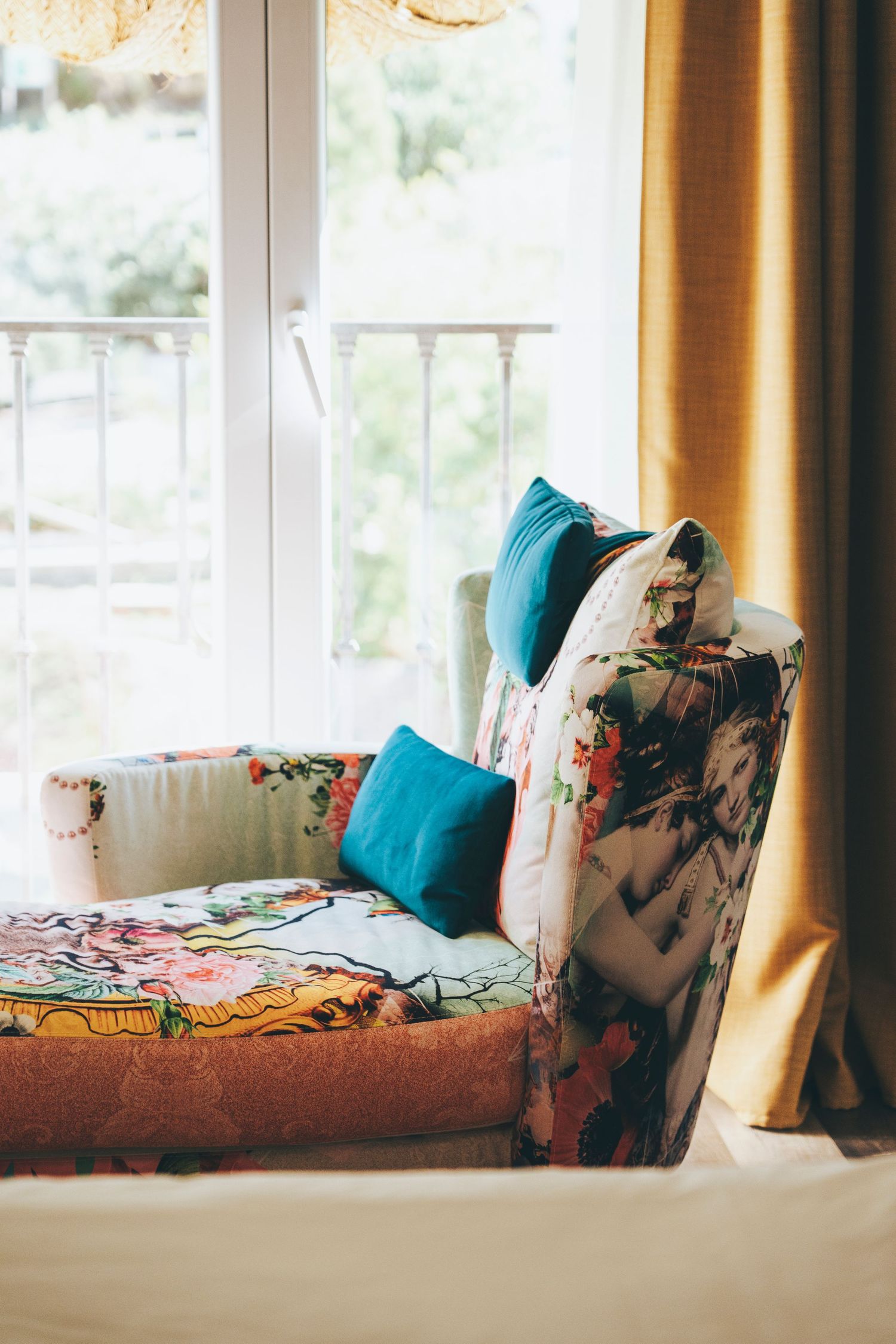 Colorful floral chaise lounge with blue cushions beside a bright window with yellow curtains in a hotel room.