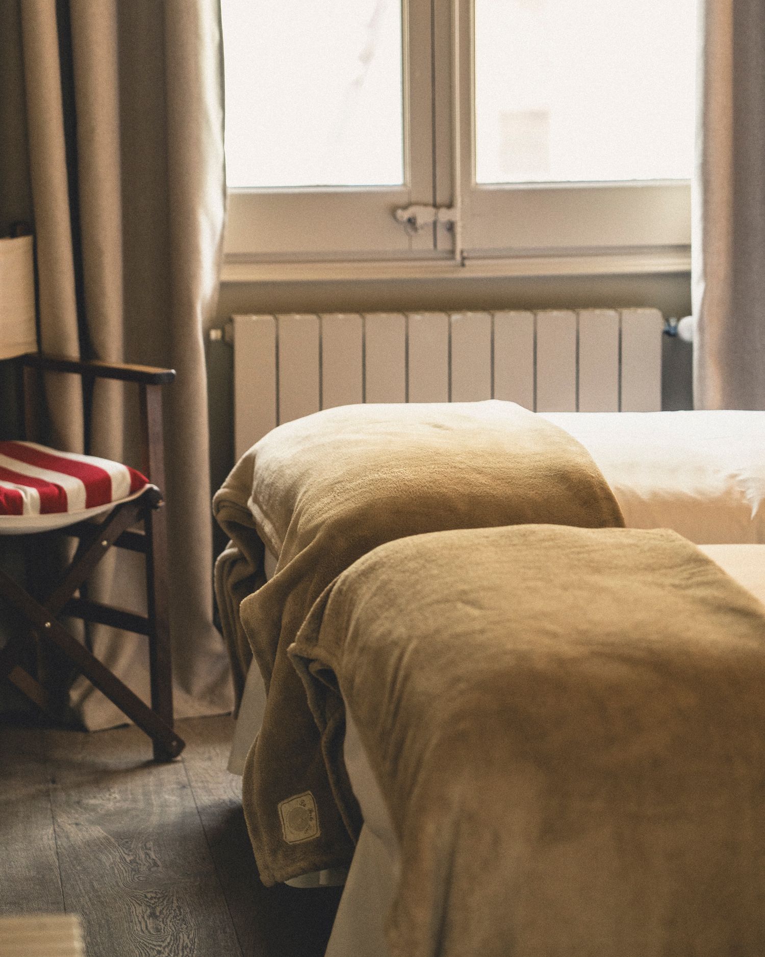 Two single beds with olive green covers near a bright window with curtains and a vintage striped chair.