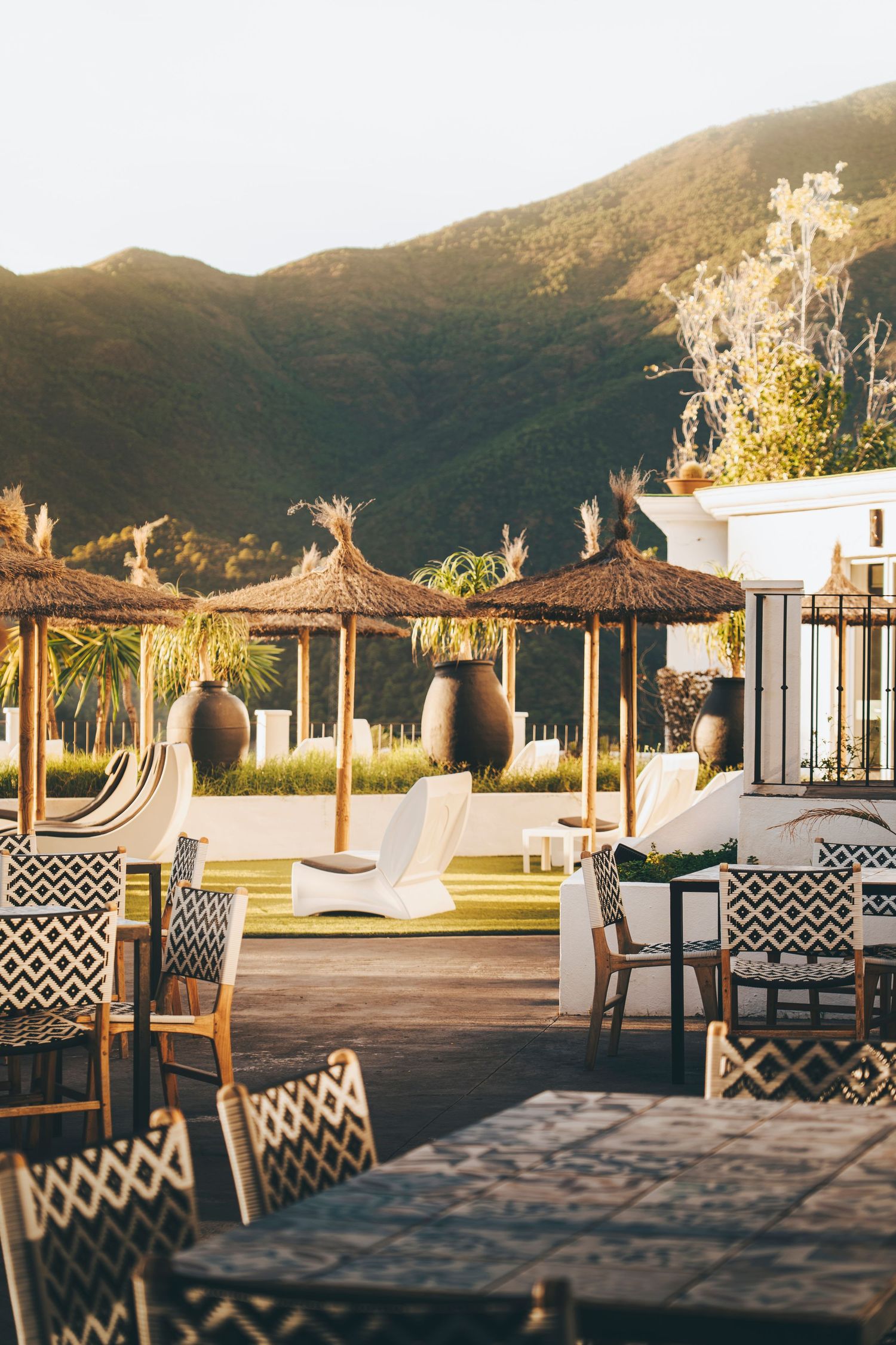 Outdoor terrace with thatched umbrellas, patterned chairs, and lush mountain backdrop in warm golden sunlight.
