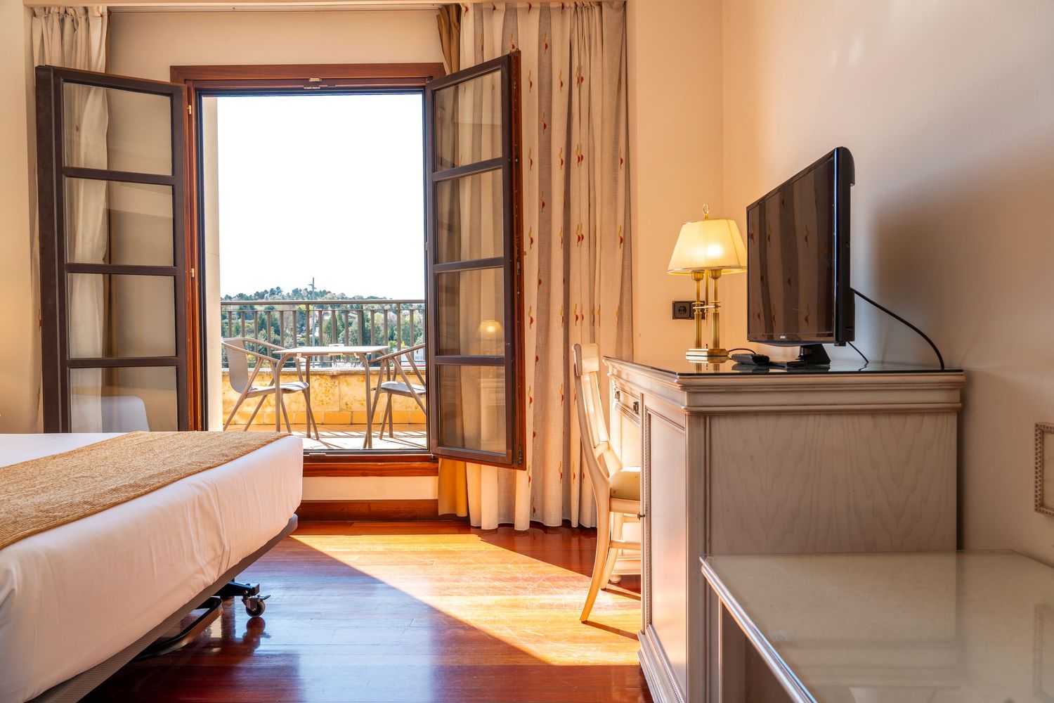 Elegant hotel room with open French balcony doors, hardwood floors, flat-screen TV, and scenic outdoor view.