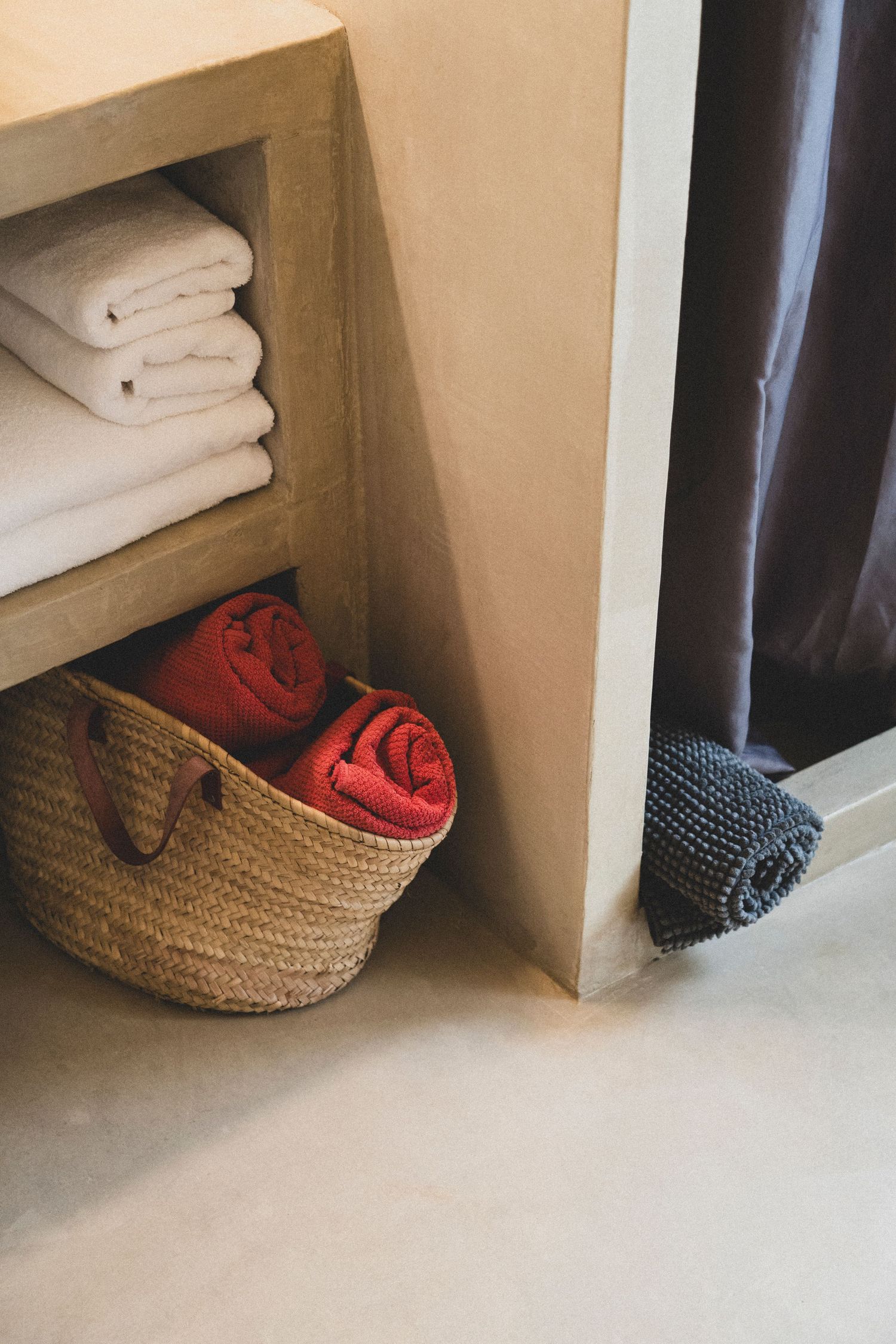 Wicker basket with rolled red towels beside a shelf with folded white towels in a minimalist bathroom.