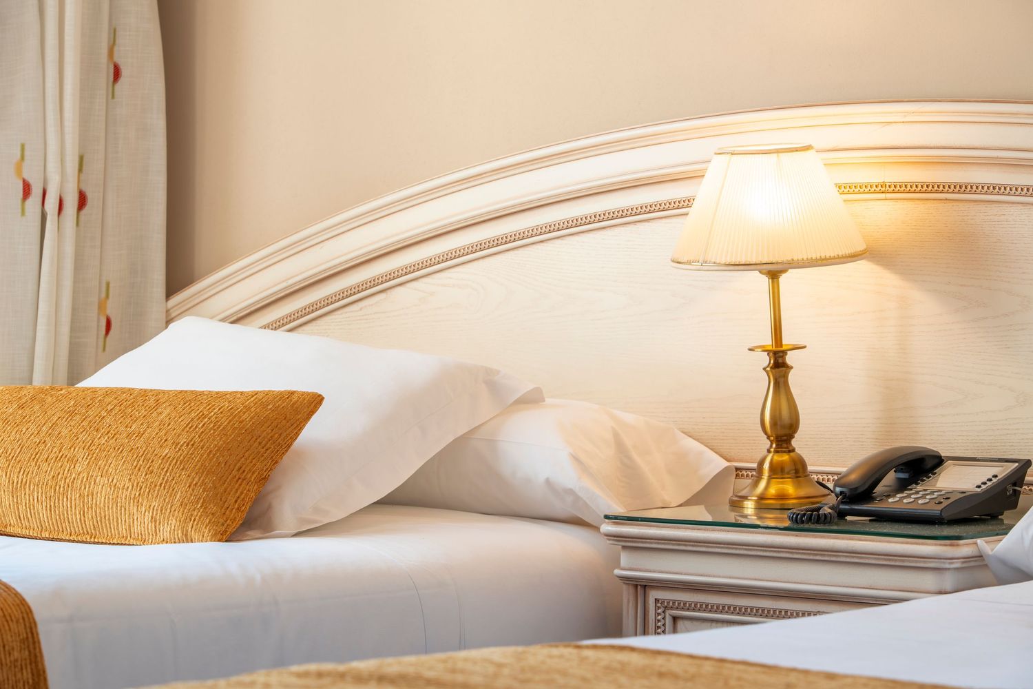 Luxury hotel twin beds with white pillows, gold lamp, ornate headboard, and telephone on bedside table.