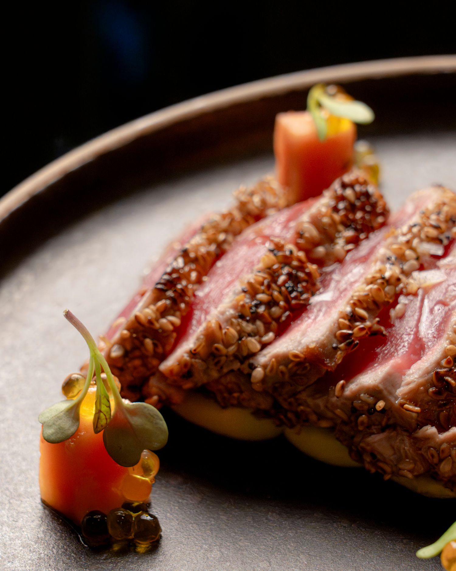Seared sesame-crusted tuna slices, rare center, plated with carrot garnish and microgreens on dark ceramic.