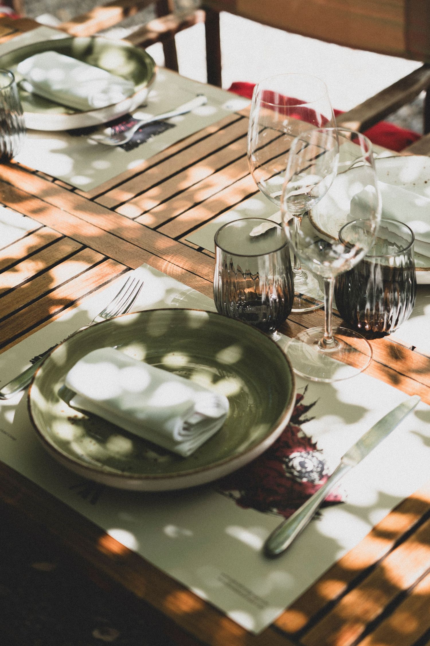 Outdoor wooden dining table set with green ceramic plates, linen napkins, and glassware in sunlight.