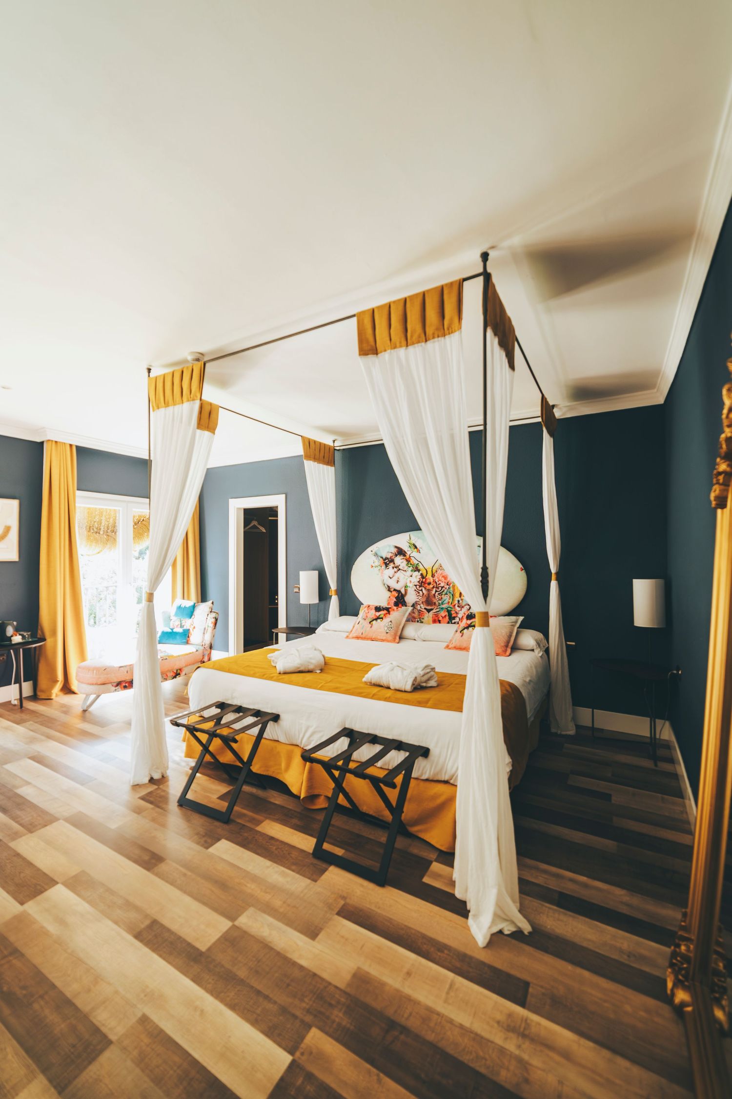 Elegant four-poster canopy bed with white drapes, yellow accents, and dark teal walls in a luxury hotel room.