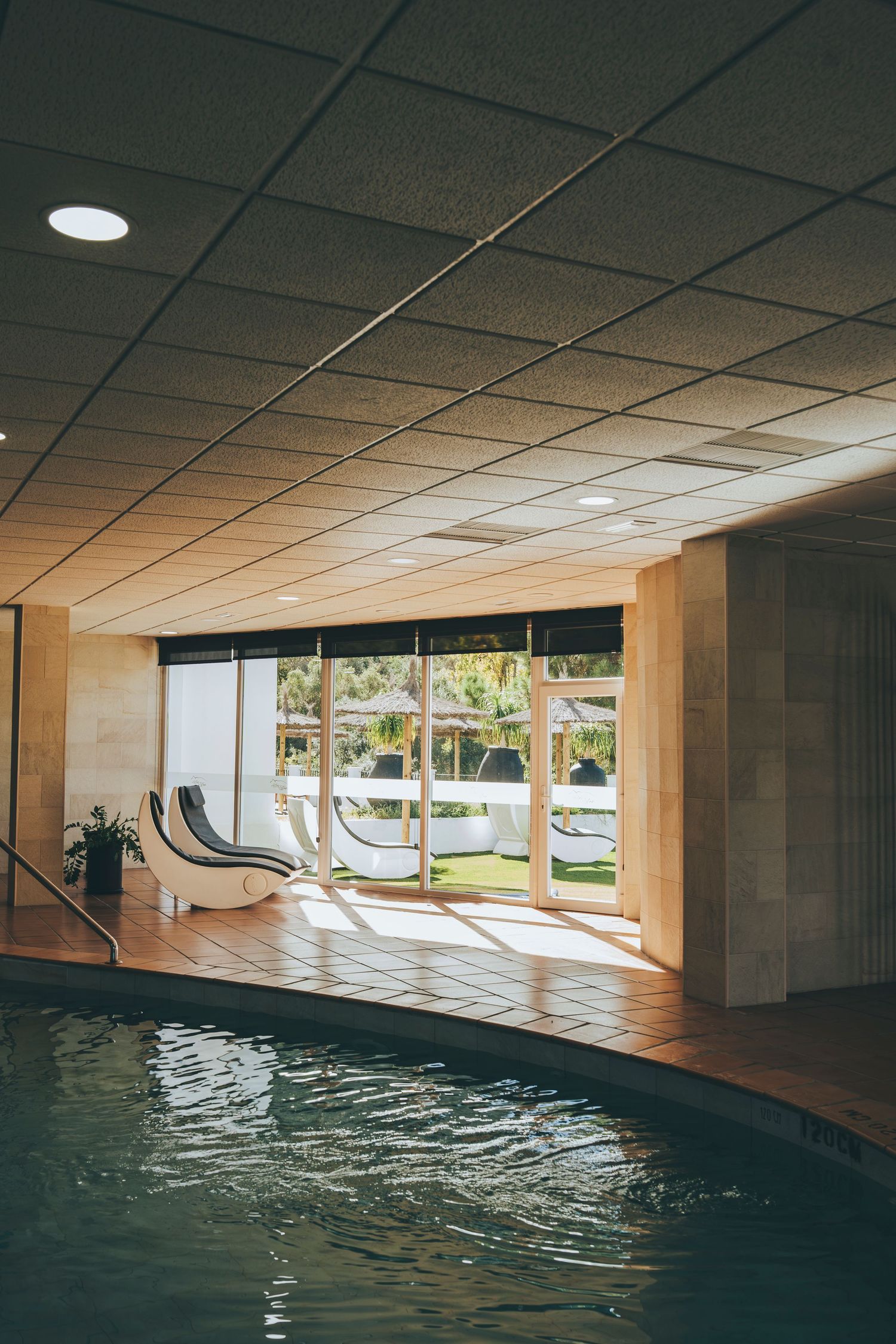 Indoor spa pool with rippling water, lounge chairs, and large glass doors opening to a sunny outdoor terrace.