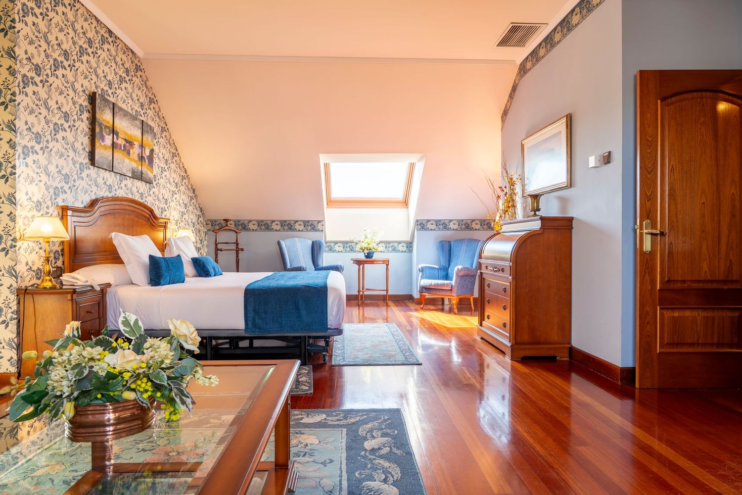 Elegant hotel room with hardwood floors, floral wallpaper, blue accents, skylight window, and classic wooden furniture.