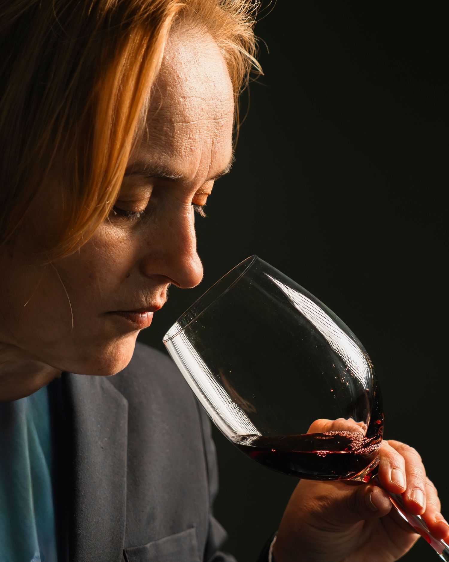 Woman with red hair smelling a glass of red wine against a dark background.