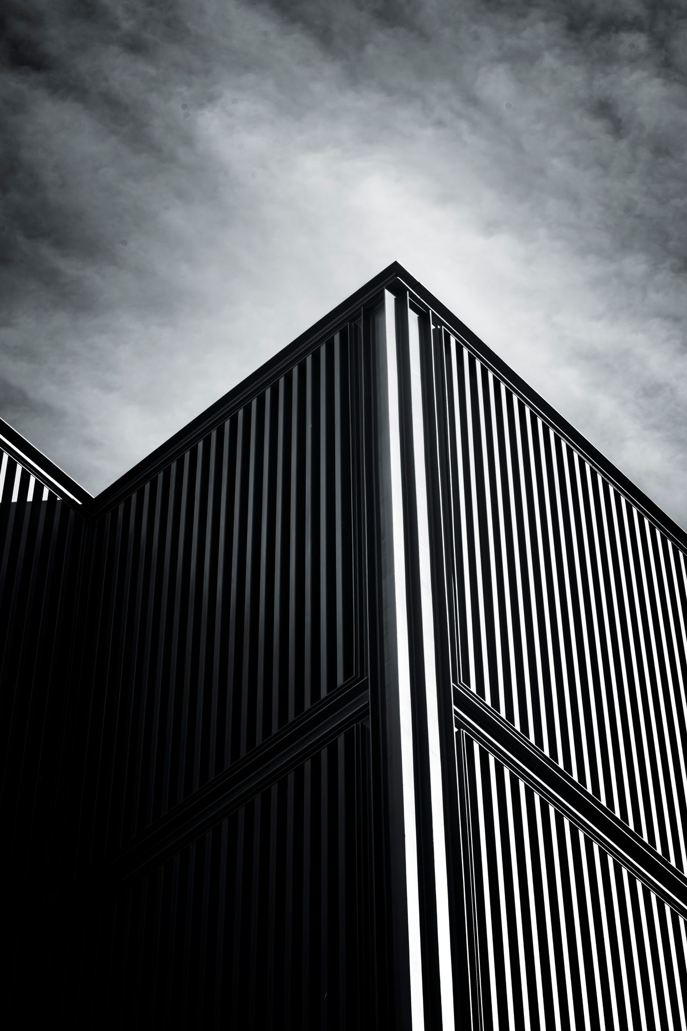 Low angle black and white photo of a modern corrugated metal building facade against cloudy sky.