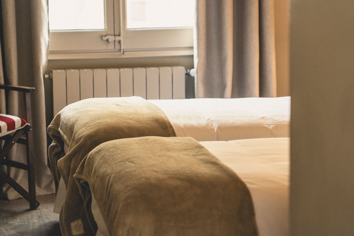 Cozy twin hotel room with two beds covered in beige blankets near window with gray curtains and radiator.