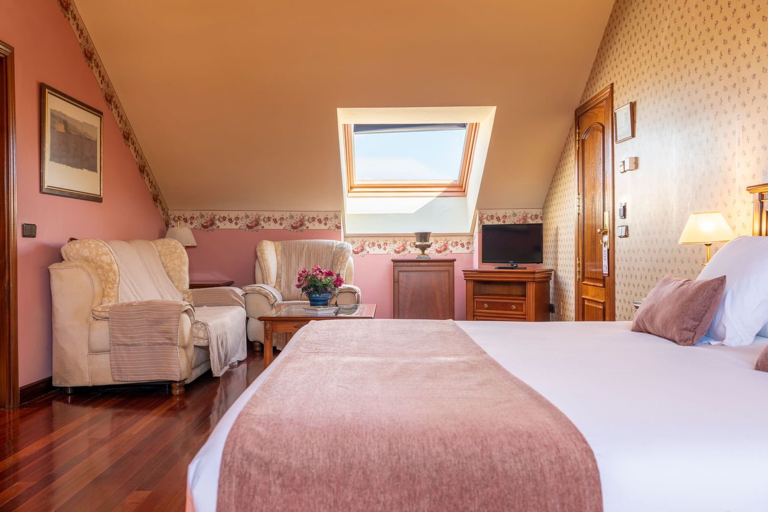 Cozy attic hotel room with skylight, pink decor, hardwood floors, double bed, and sitting area with armchair.