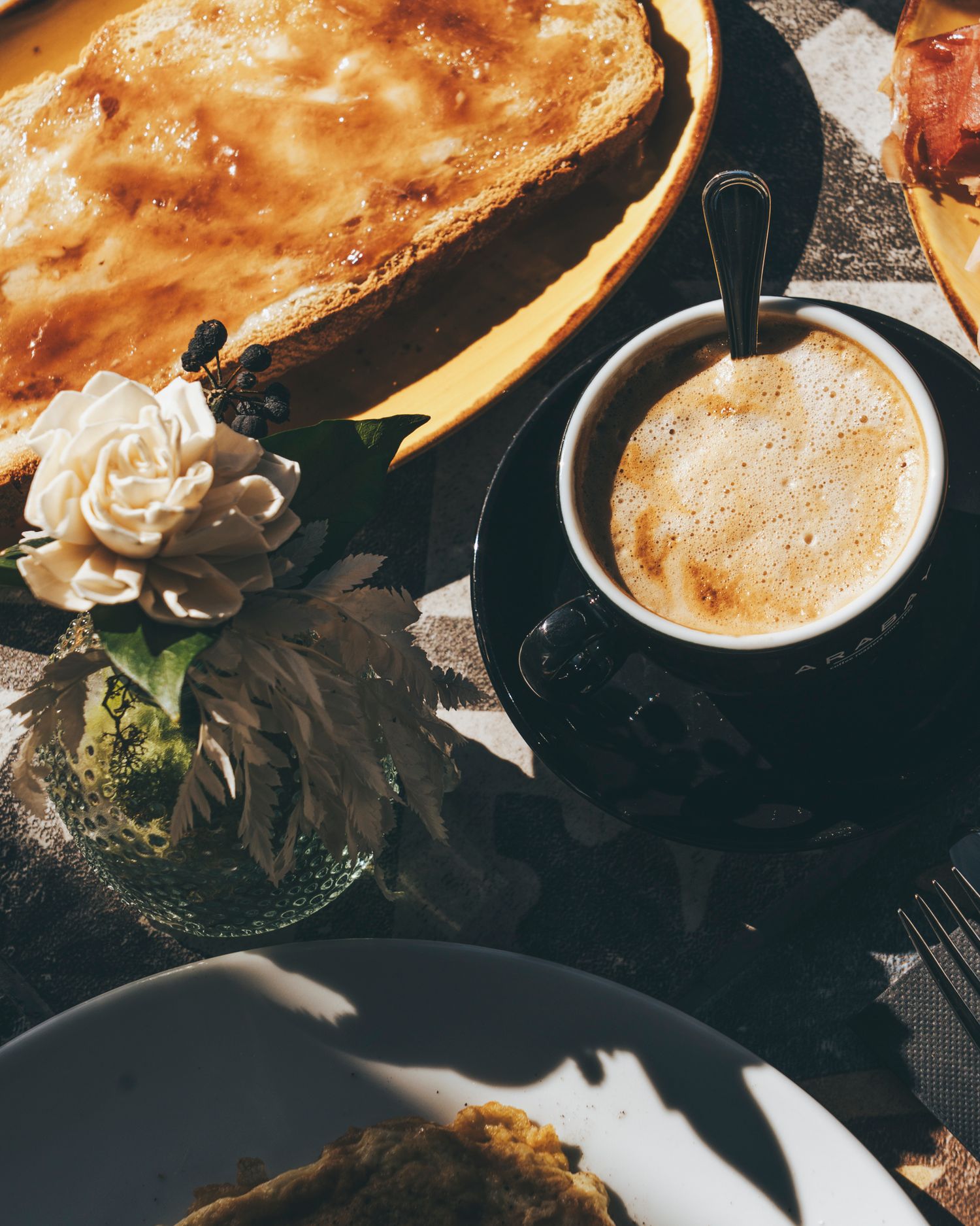 Frothy espresso coffee beside a golden baked tart in warm sunlight on a cafe table.