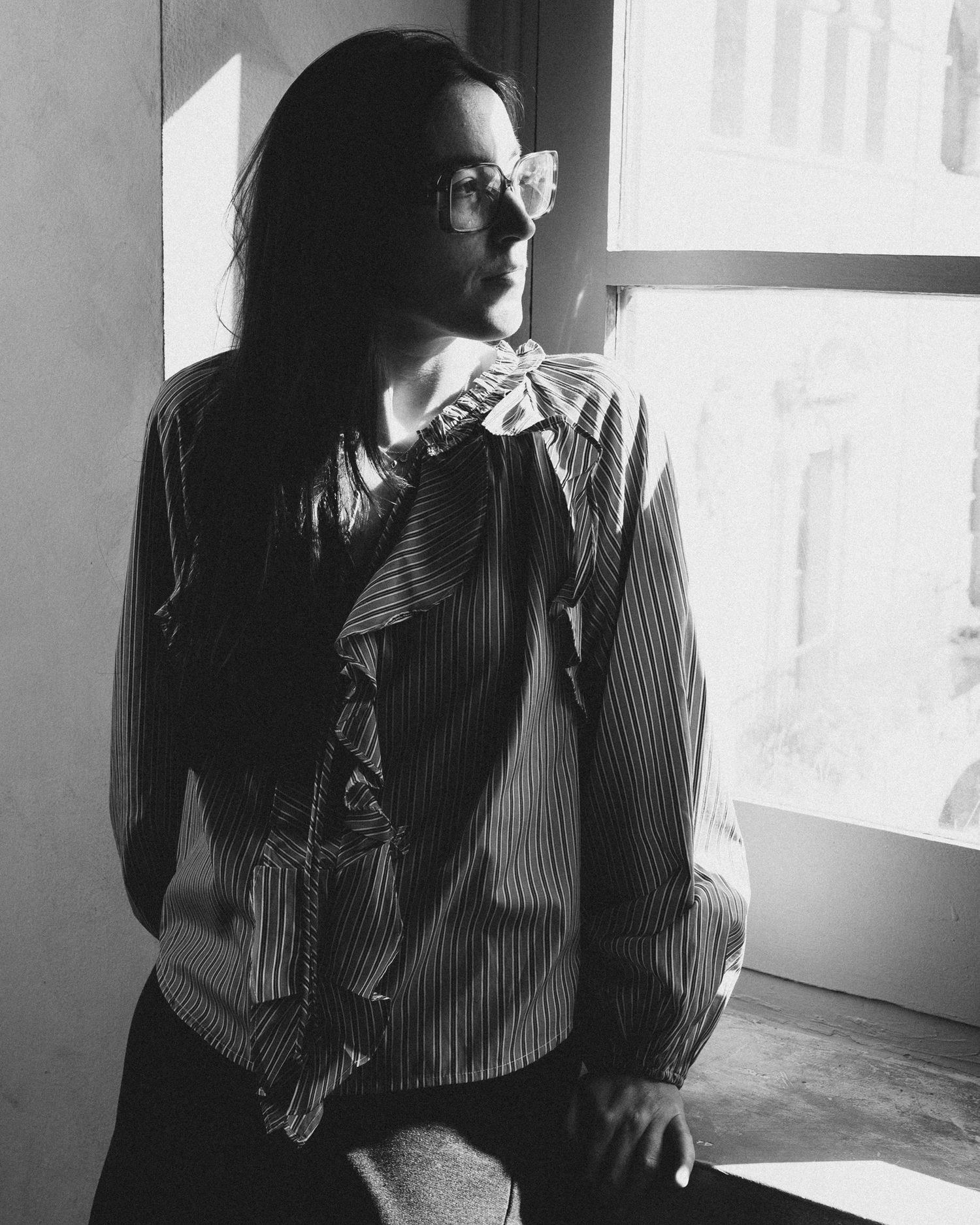 A person with long hair and glasses poses by a window in a striped ruffled blouse in black and white.