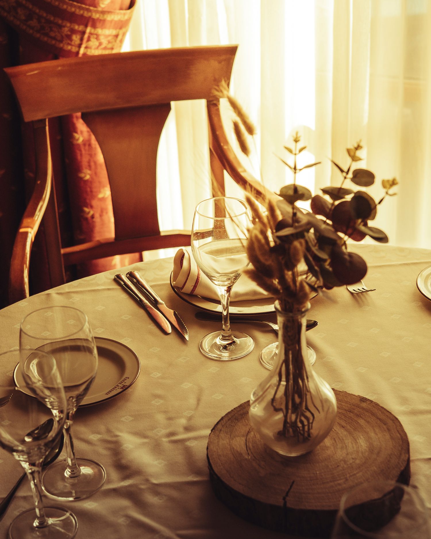 Warmly lit restaurant table set with wine glasses, plates, and dried botanical centerpiece in glass vase on wood slice.