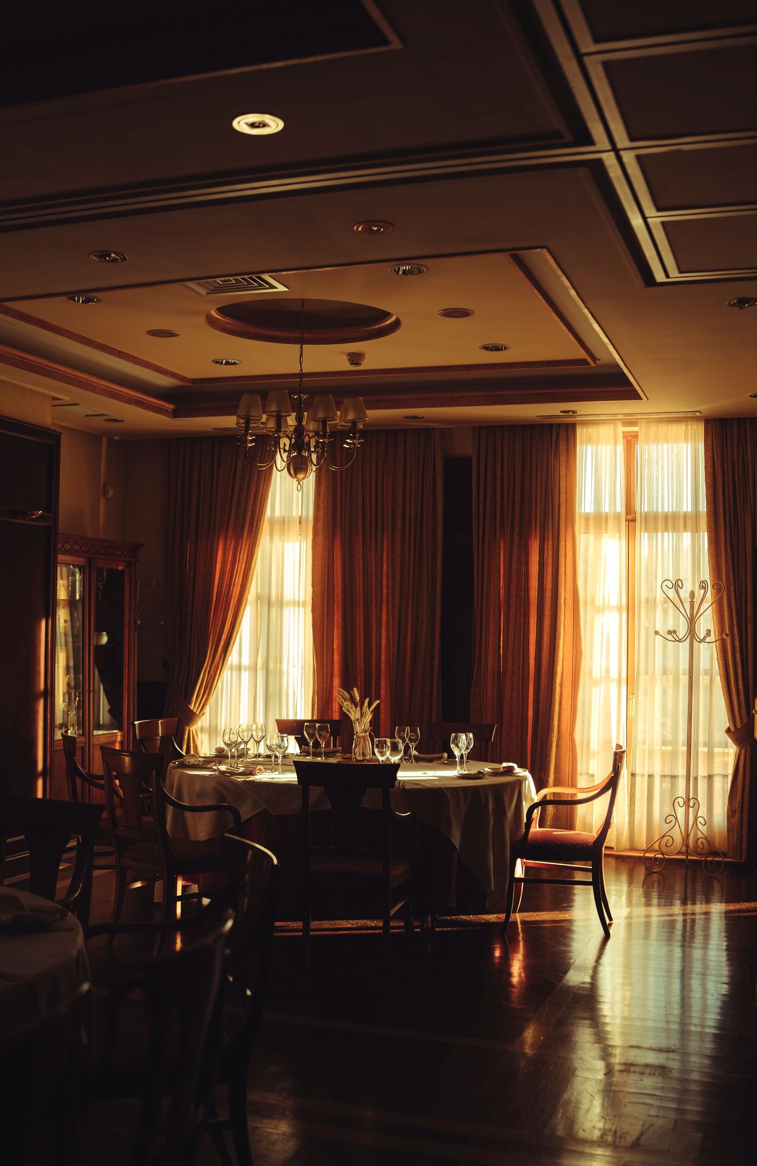 Warmly lit fine dining room with round tables, wine glasses, chandelier, and golden draped windows.