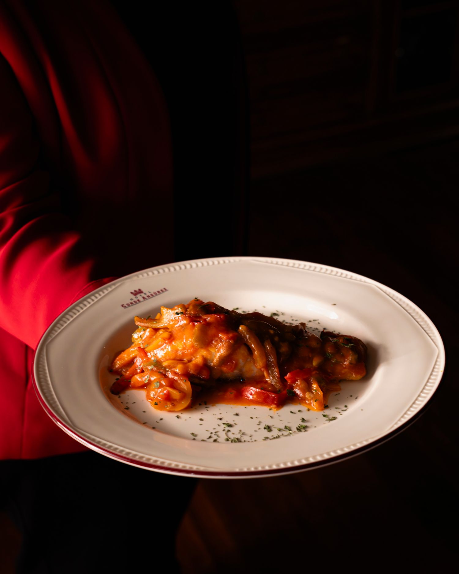 A server presenting a plate of chicken in tomato sauce on a white restaurant plate.