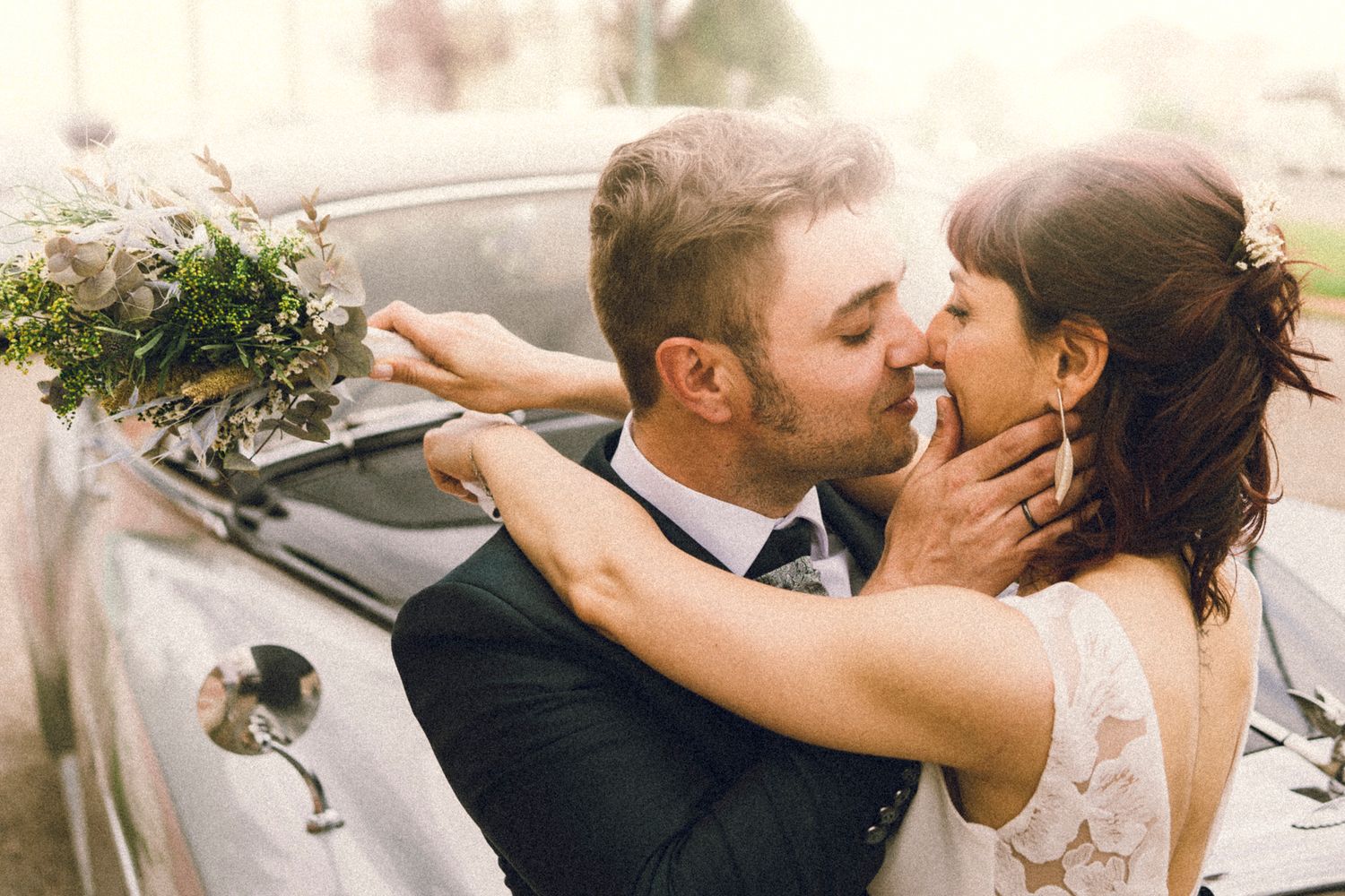 Newlyweds kissing tenderly near a vintage car, bride holding a lush green and white bouquet.