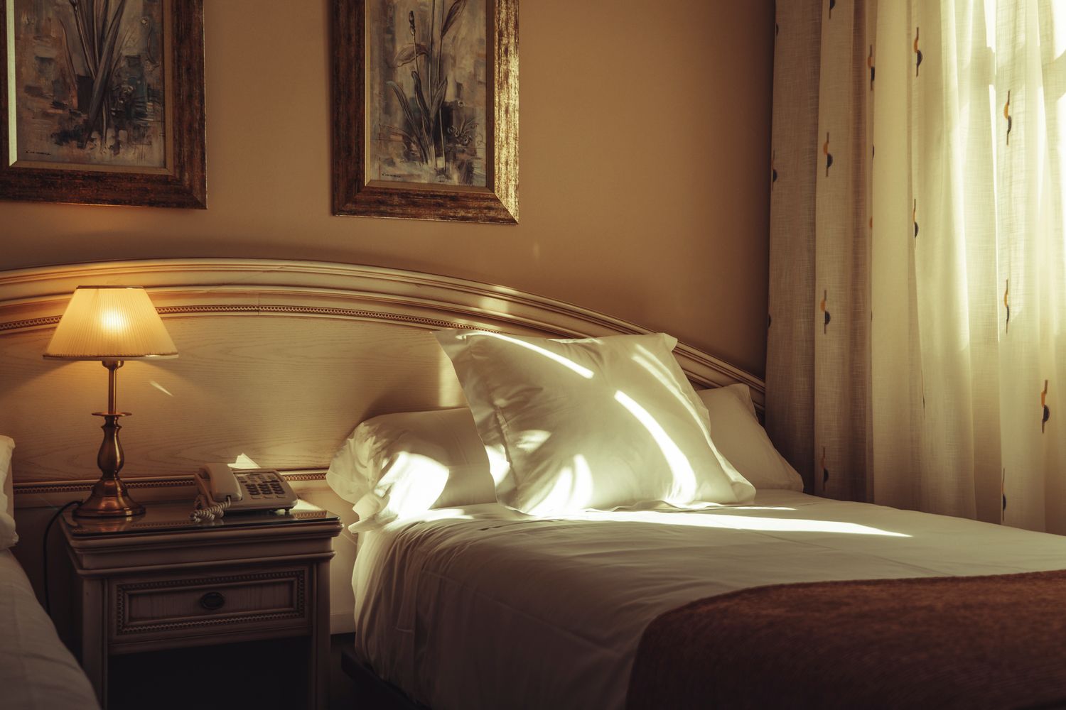 Elegant hotel bedroom with warm lamp light, white pillows on a wooden headboard bed, and sunlight streaming through sheer curtains.