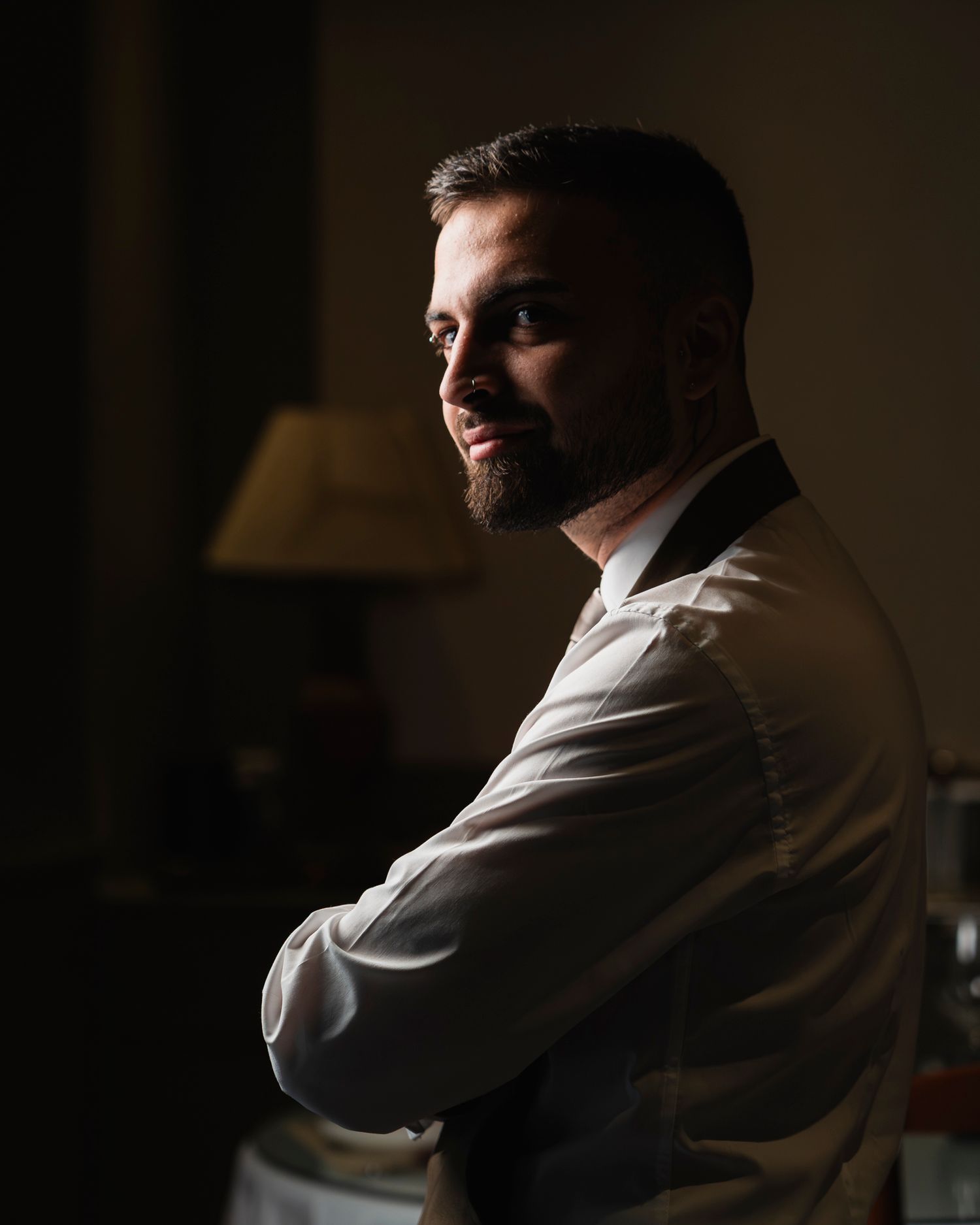 A man with a beard stands with arms crossed in dramatic side lighting wearing a white dress shirt.