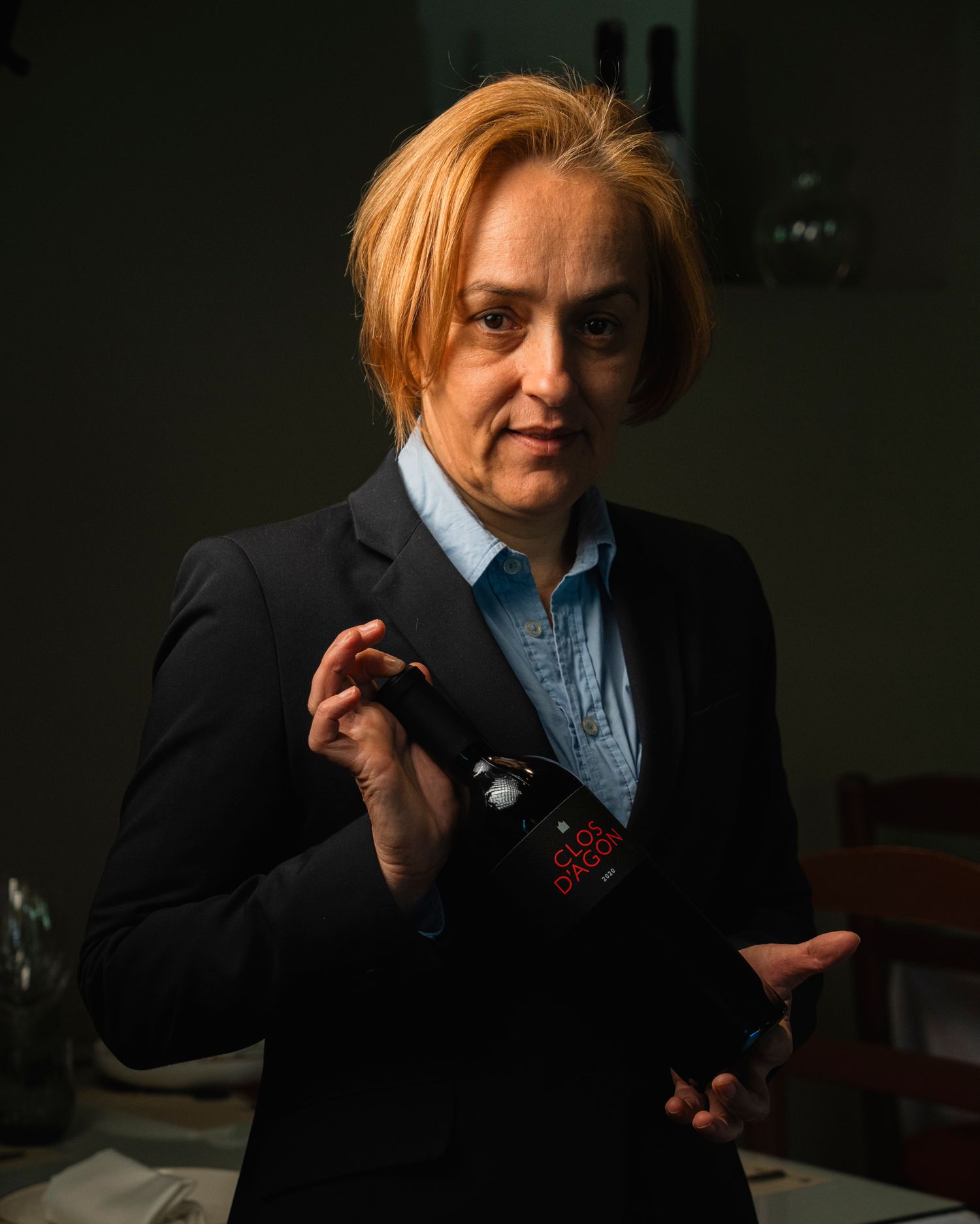 A woman with red hair in a black blazer holds a camera in a dimly lit room.